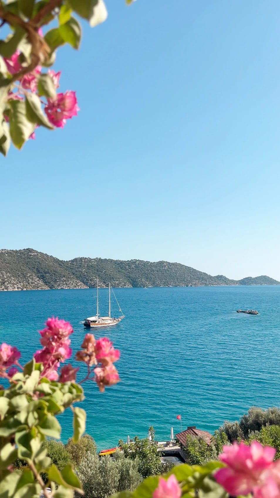 Pinke Bougainvillea rahmt den Blick auf eine Gulet in einer türkisblauen Bucht.