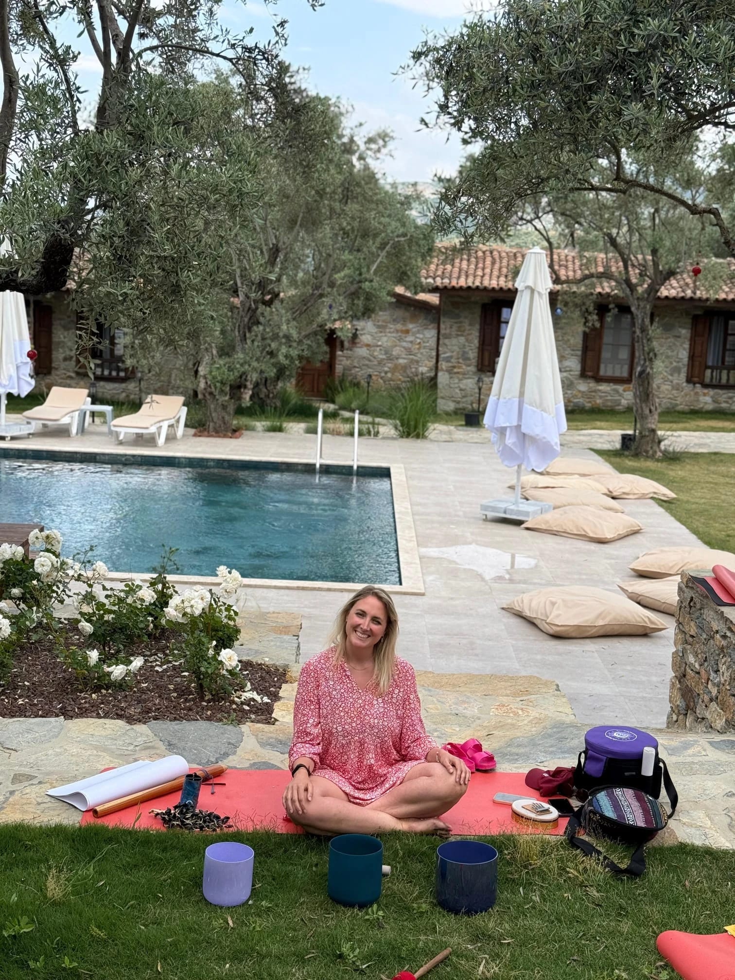 Eine Frau sitzt lächelnd auf einer Matte vor dem Pool, vor ihr stehen Kristallklangschalen.