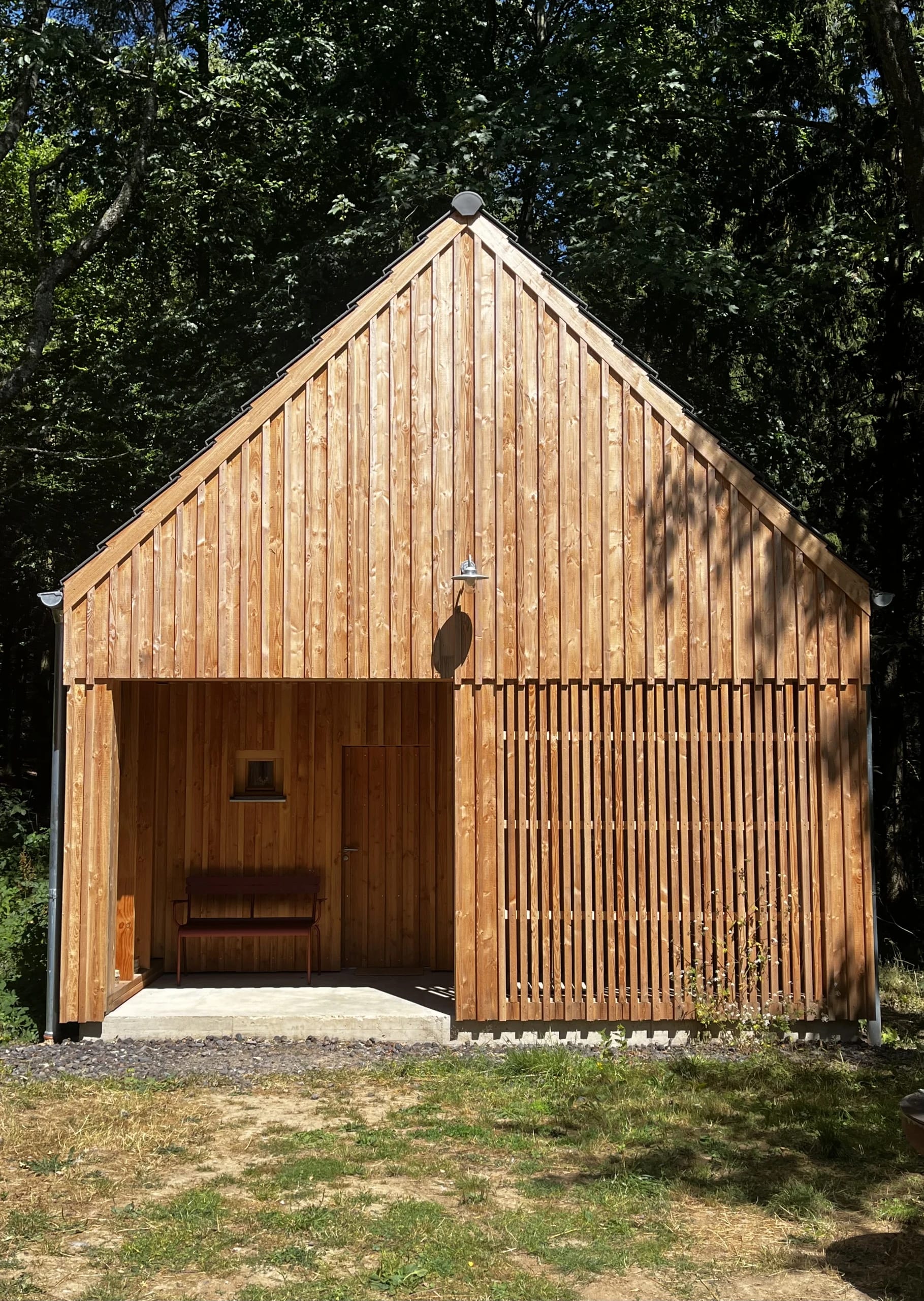 Die Saunahütte aus hellem Holz am Waldrand, davor eine überdachte Terrasse mit Bank und eine Außendusche.