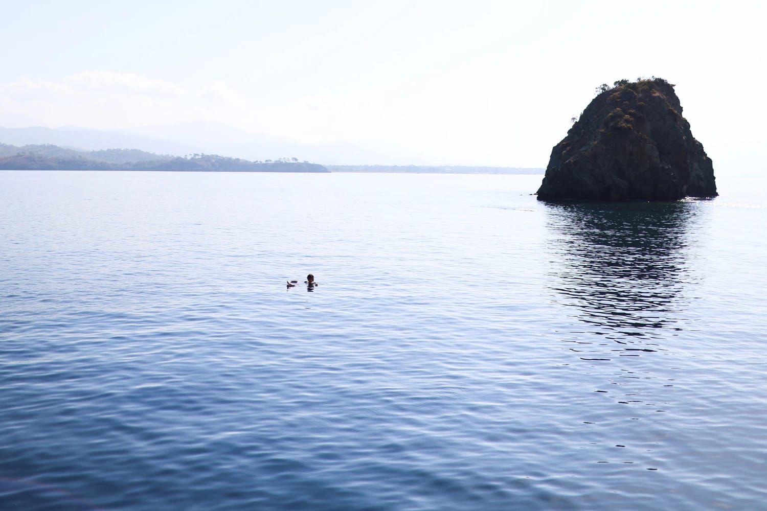 Eine Schwimmerin im weiten, ruhigen Meer neben einem markanten Felsen