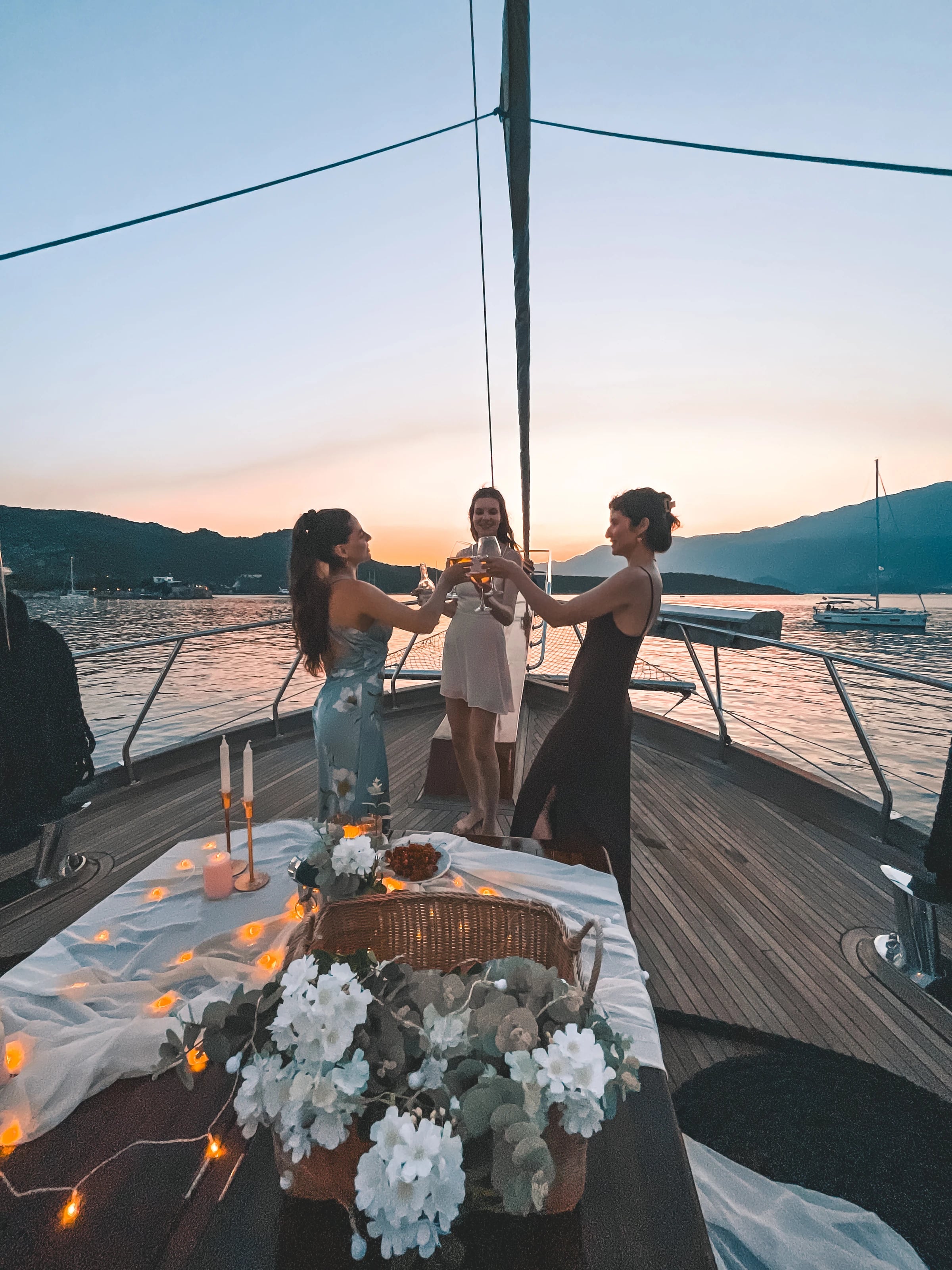Drei Freundinnen stoßen am Bug der Yacht mit Gläsern an, daneben ein liebevoll dekorierter Tisch mit Lichterkette im Abendrot.