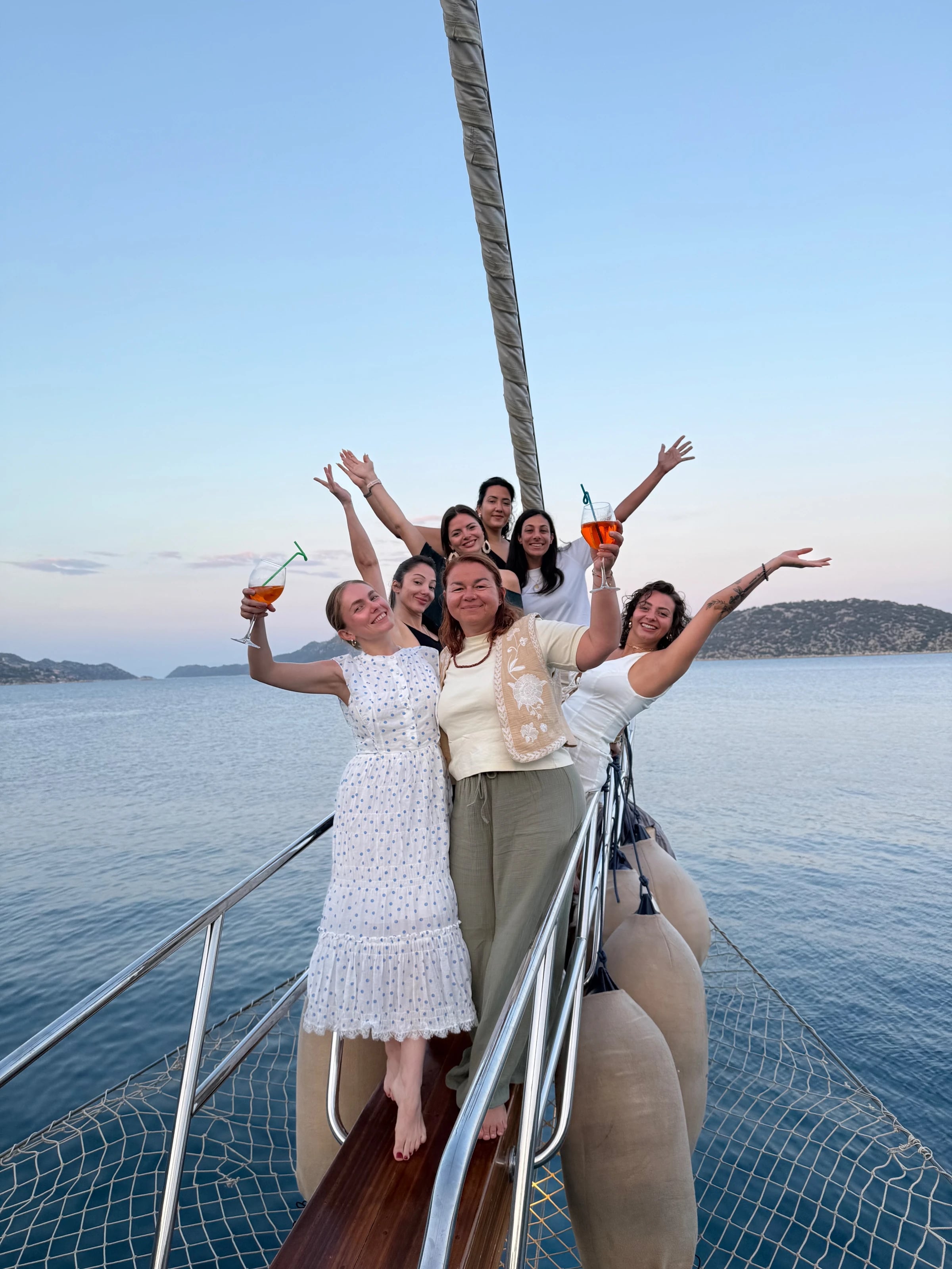 Eine Gruppe lachender Frauen steht mit Spritz-Gläsern und erhobenen Armen am Bug der Yacht.