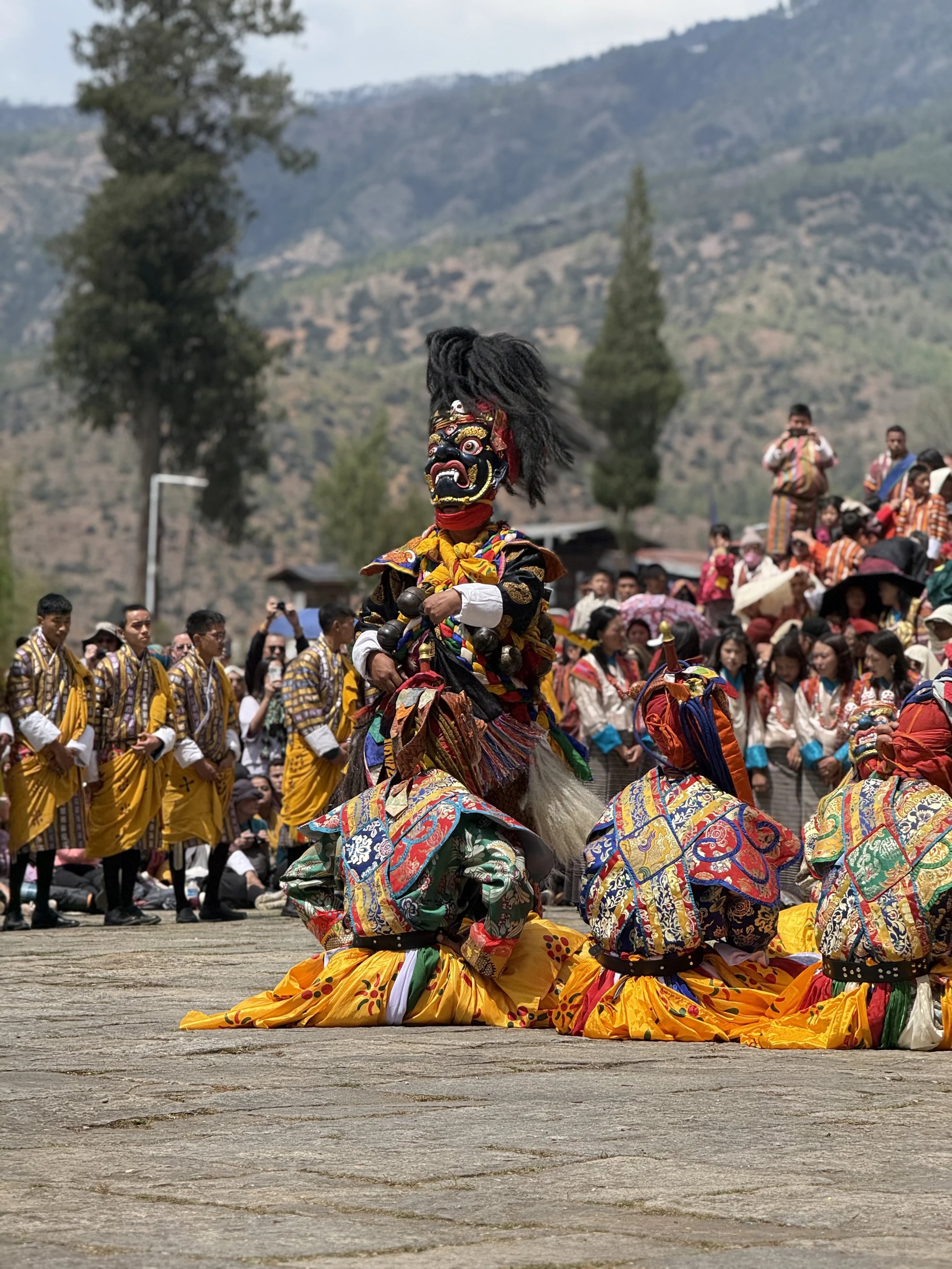 Maskierte Cham-Tänzer bewegen sich im Innenhof des Dzong, umringt von Zuschauern in traditioneller Kleidung.