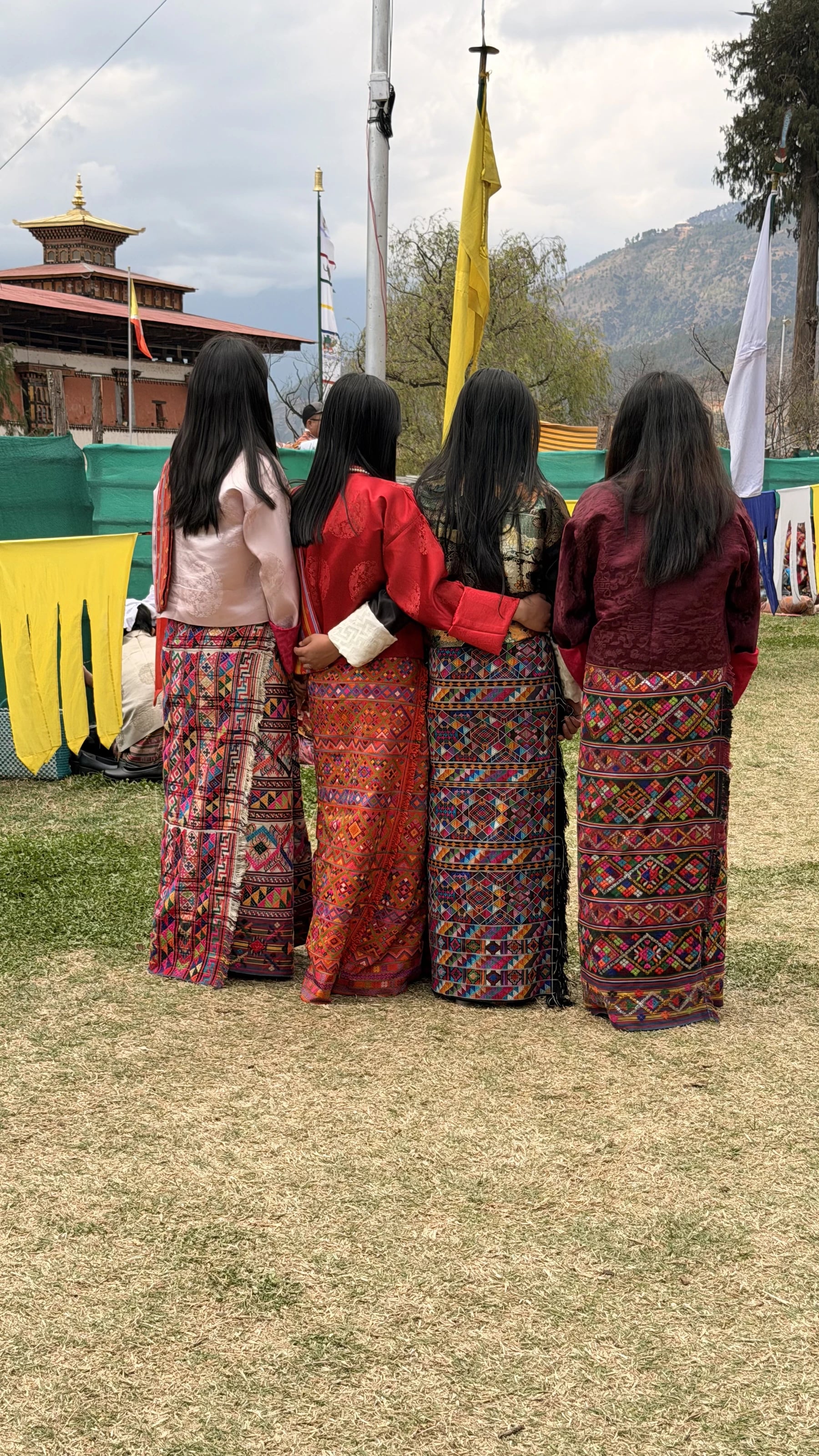 Vier Frauen in traditioneller bhutanischer Tracht (Kira) stehen mit dem Rücken zur Kamera bei einem Fest.