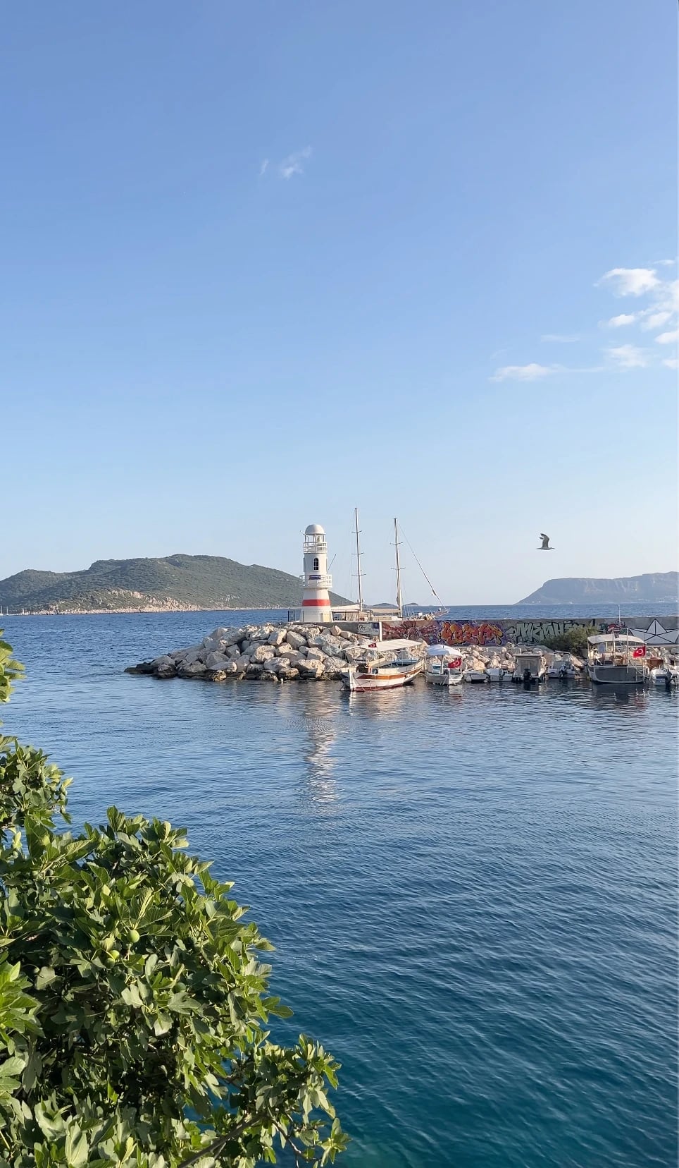 Der kleine Leuchtturm an der Hafenmole von Kaş mit Fischerbooten im ruhigen blauen Wasser.