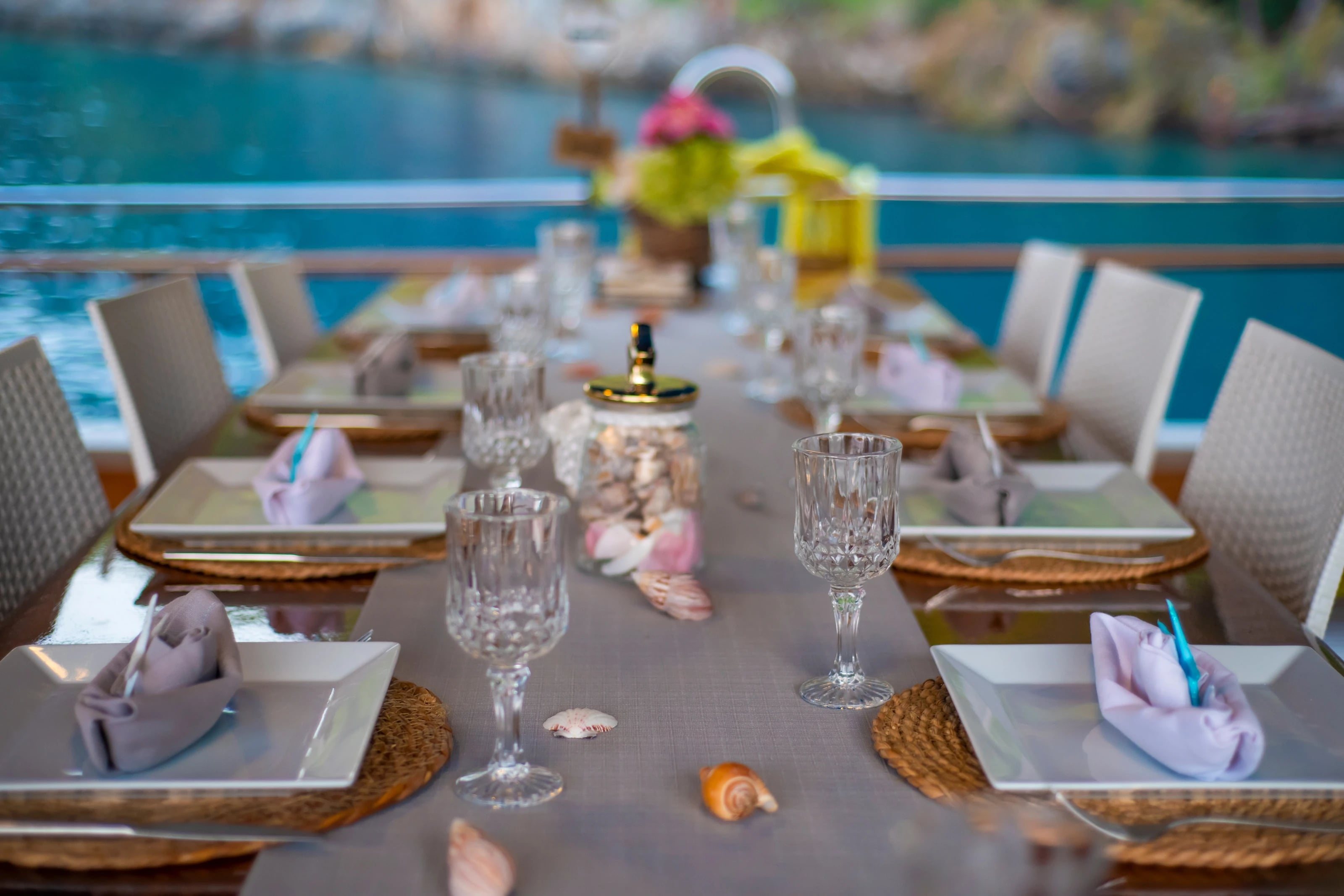 Festlich gedeckter langer Tisch an Deck mit Kristallgläsern, Muscheln und Blick auf das türkisfarbene Wasser.