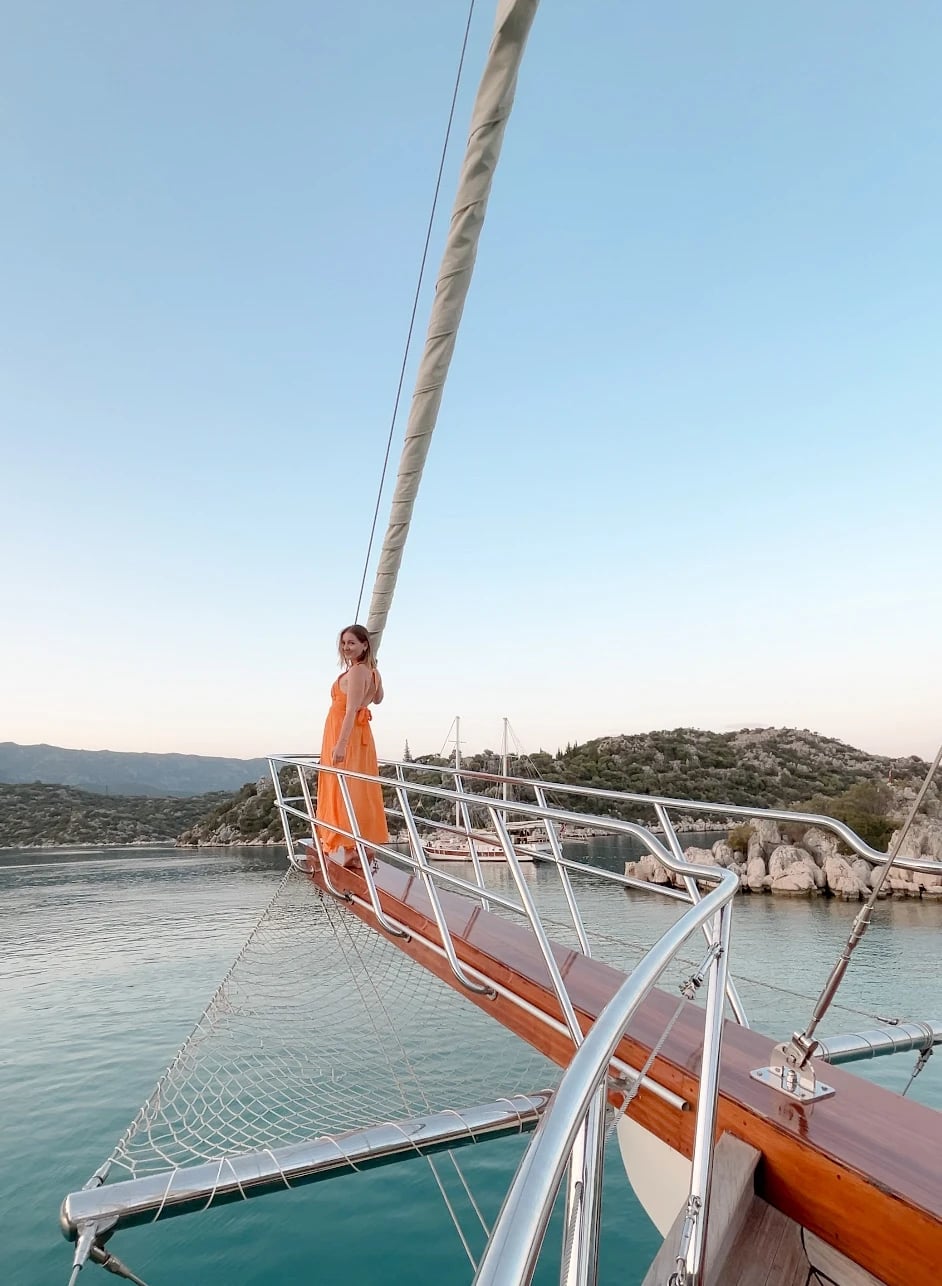 Saskia im orangefarbenen Kleid steht am Bug einer Gulet, dahinter eine ruhige Bucht im Abendlicht.