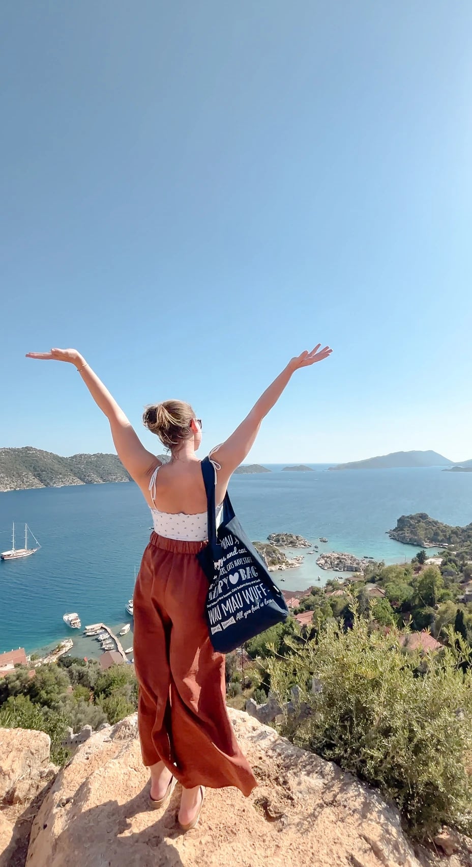 Eine Frau breitet auf einem Felsen hoch über der Bucht von Kaleköy die Arme aus, unten liegen Boote und kleine Inseln.