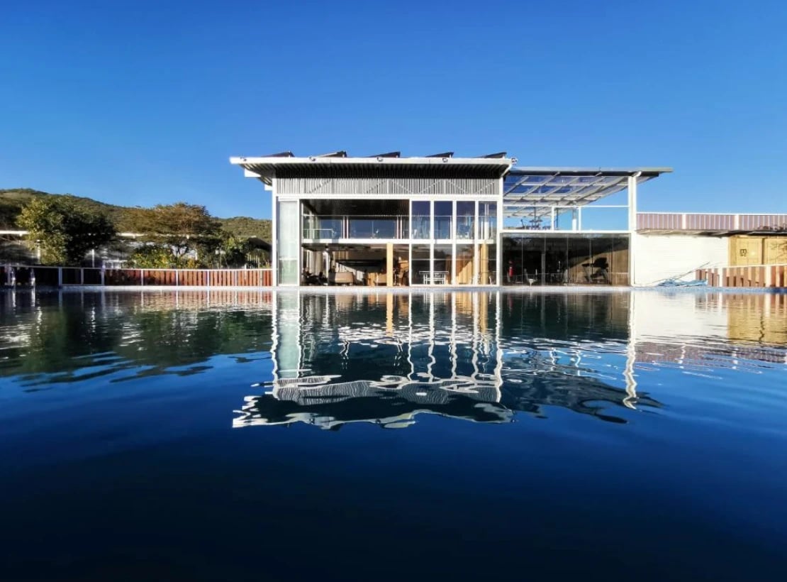 Das gläserne Haupthaus der Lodge spiegelt sich im Pool unter tiefblauem Himmel.