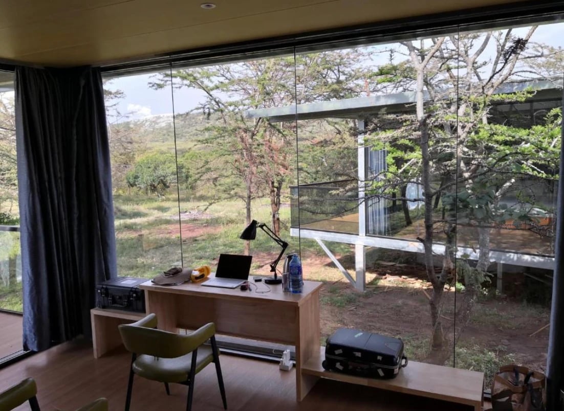 Lodgezimmer mit Schreibtisch vor einer großen Glasfront mit Blick auf Akazien und Buschland.
