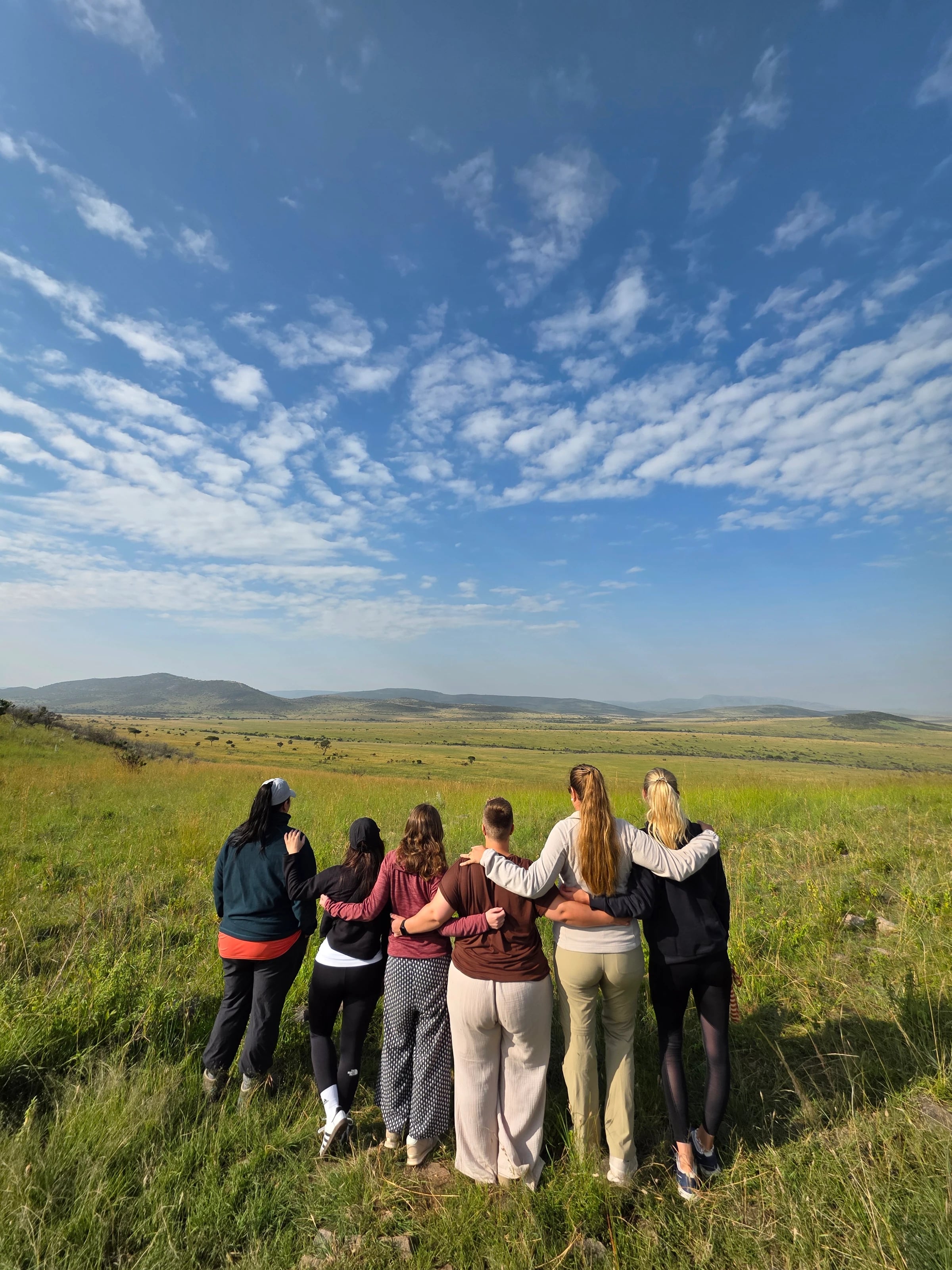 Sechs Frauen stehen Arm in Arm im hohen Gras und blicken über die weite Savanne unter blauem Himmel.