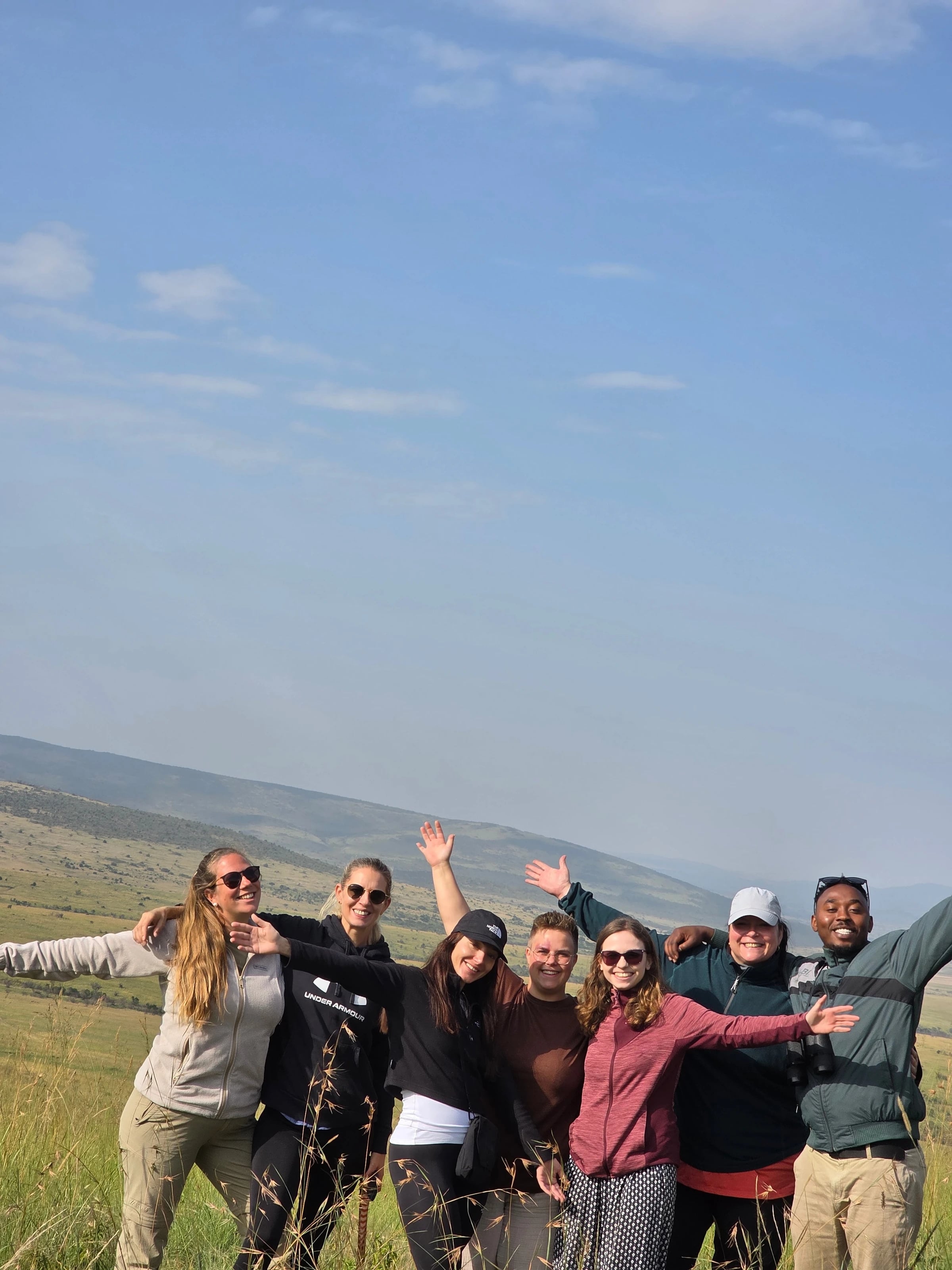 Eine fröhliche Reisegruppe steht Arm in Arm im Grasland und reckt die Hände in den Himmel.