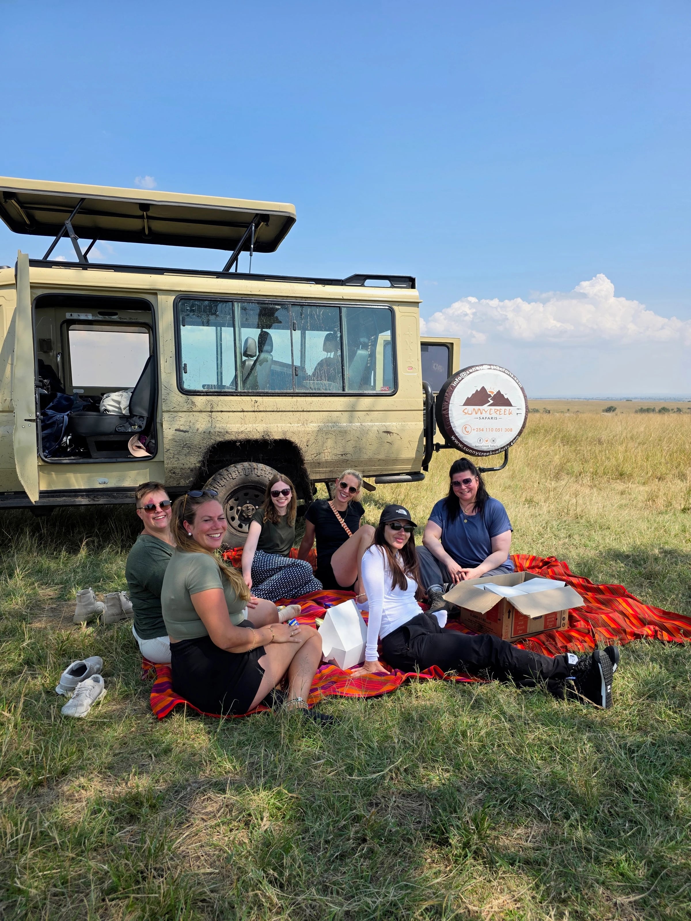 Frauen machen Picknick auf einer bunten Decke im Gras neben einem Safarijeep.