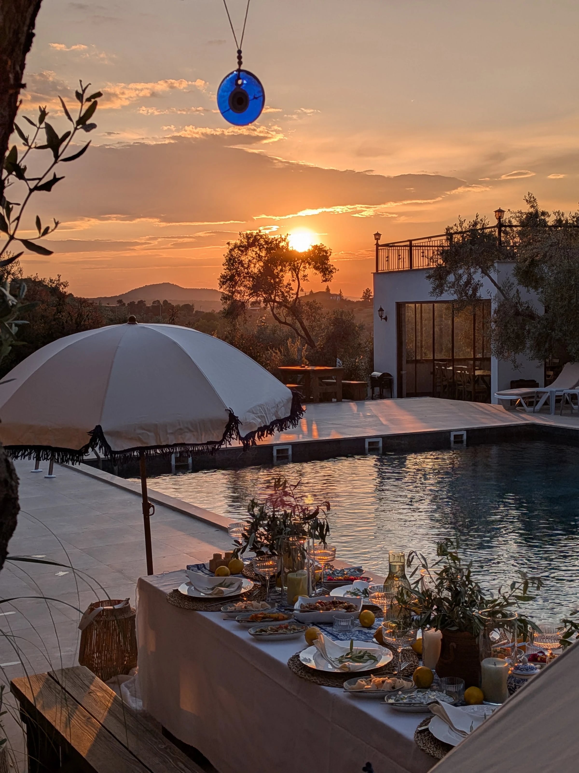 Gedeckte Tafel am Pool im Sonnenuntergang, darüber ein Sonnenschirm und ein blaues Nazar-Auge.