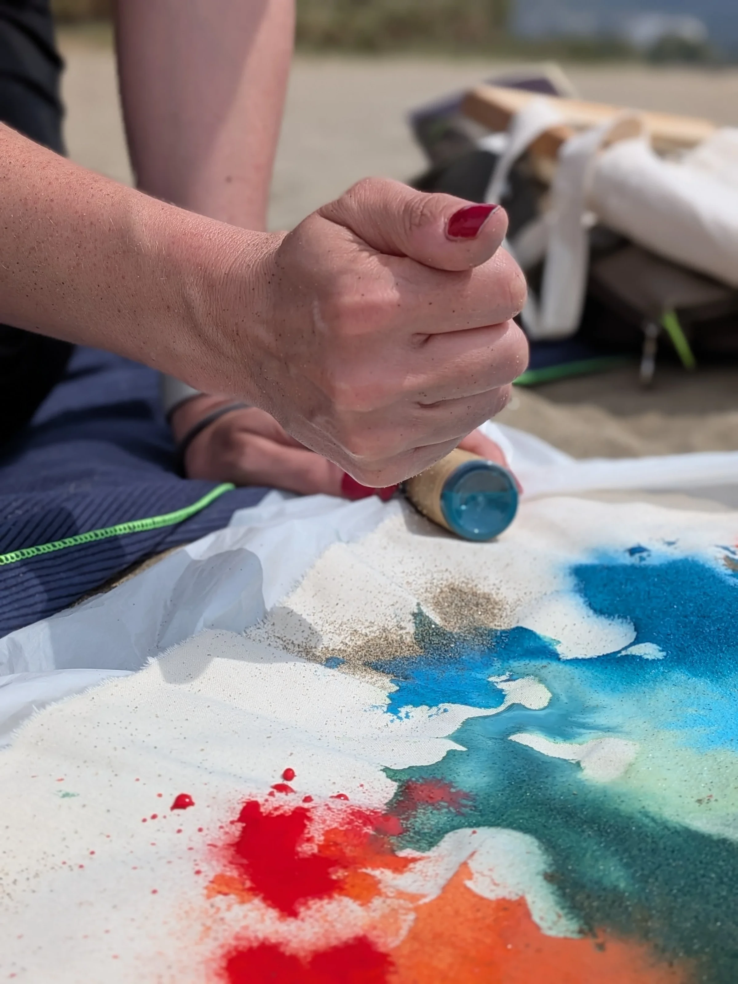 Hände rollen mit einer Farbrolle Blau und Rot über ein weißes Blatt im Sand.