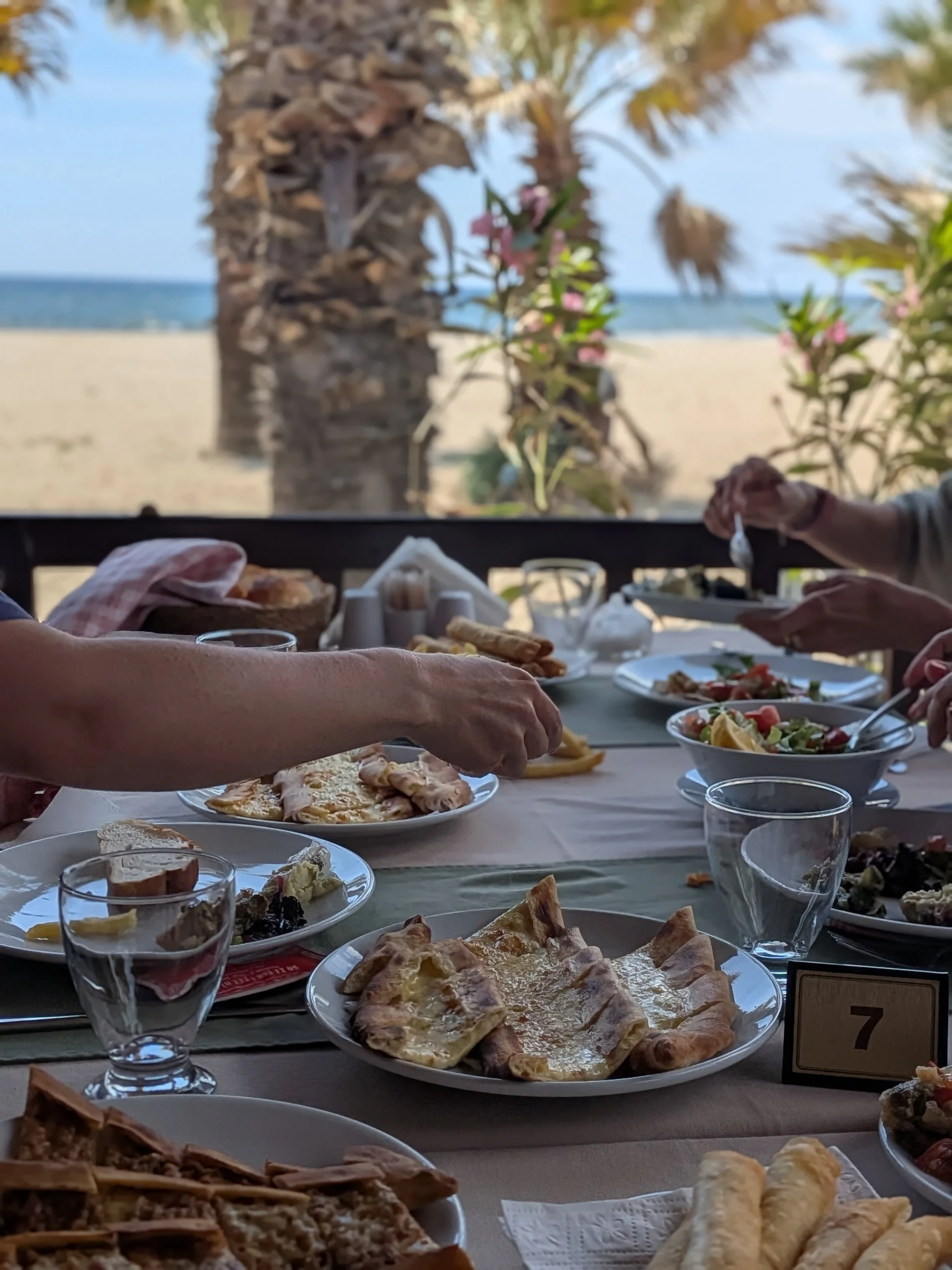 Gedeckter Tisch mit Mezze und Brot, dahinter Palmen und der Strand am Meer.