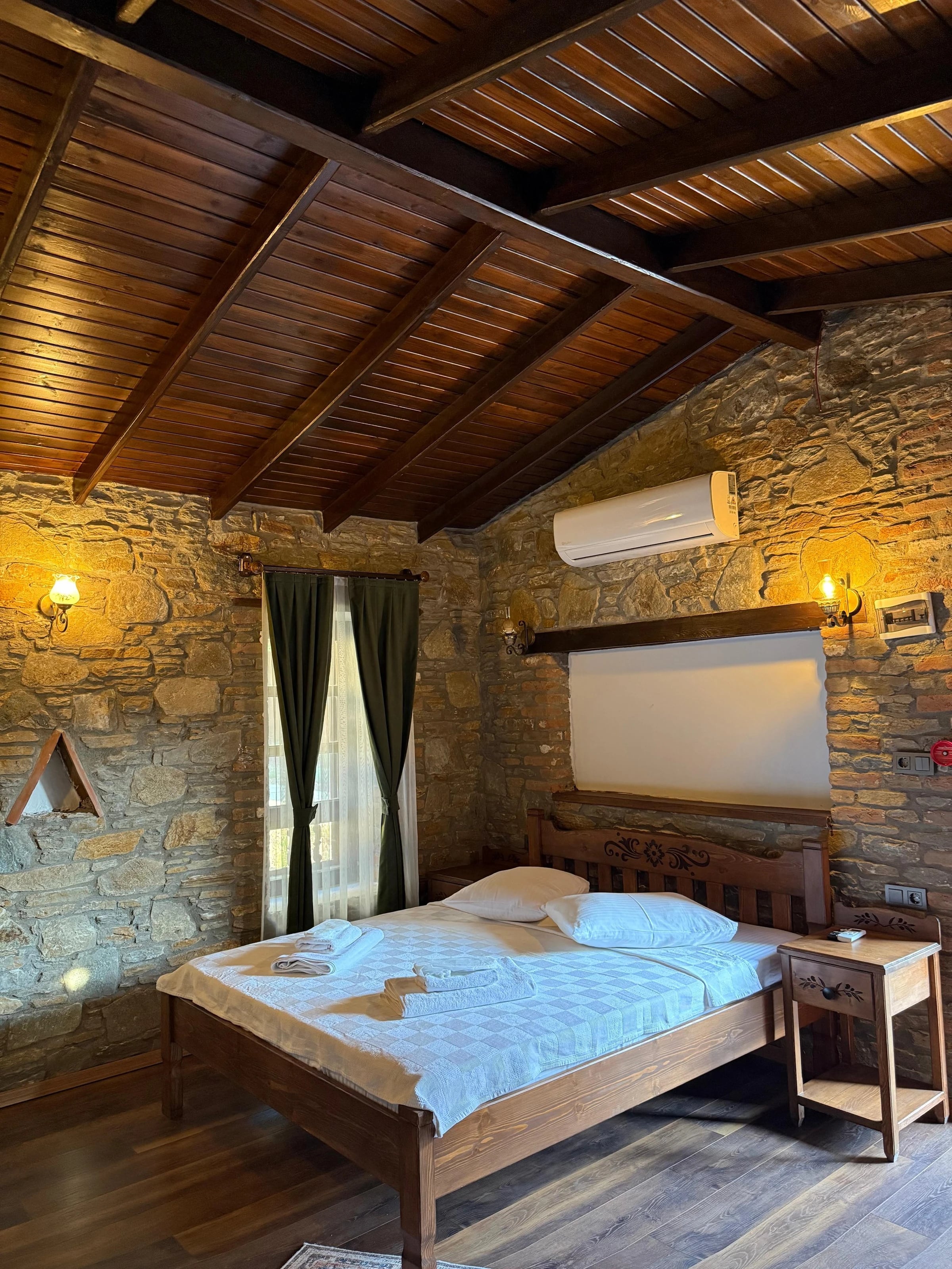 Schlafzimmer mit Natursteinwand, Holzdecke, Doppelbett und Fenster mit Vorhang.