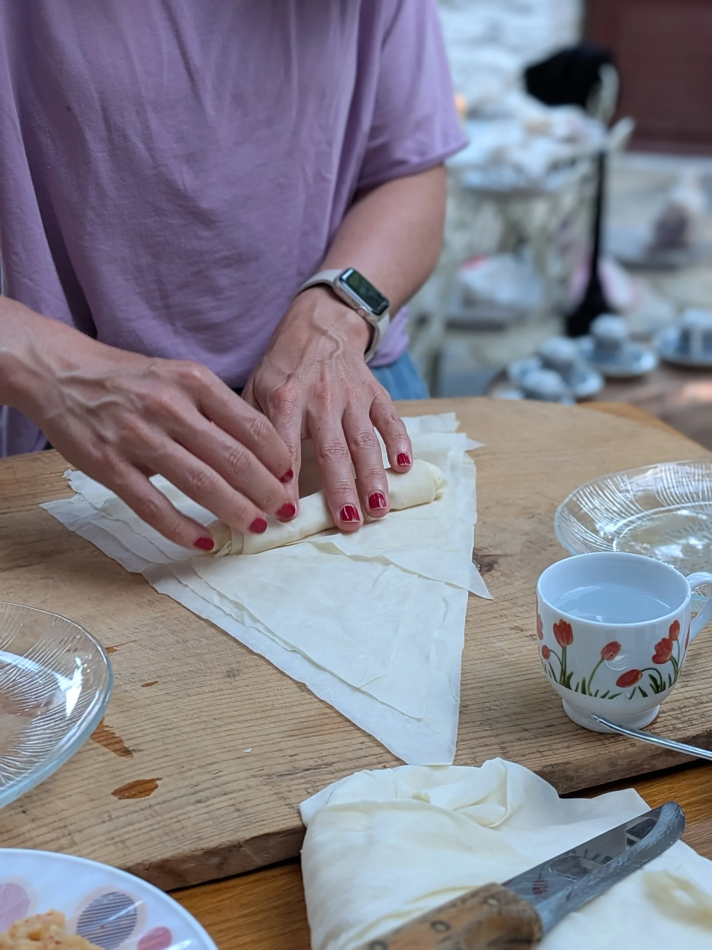 Hände falten auf einem Holztisch dünnen Teig zu Taschen, daneben steht eine Tasse mit Mohnmuster.