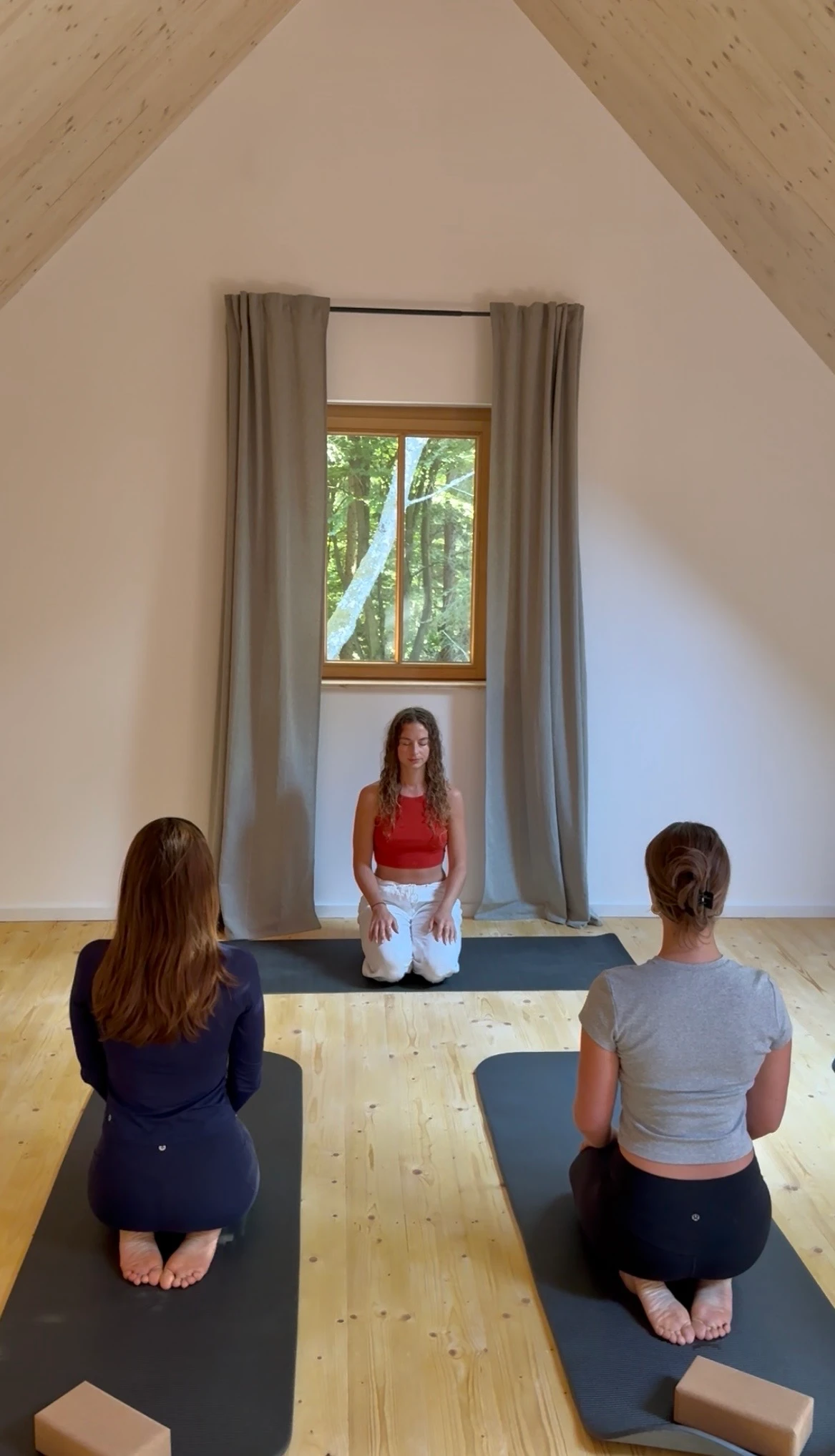 Frauen meditieren im Yogaraum, die Leiterin sitzt ihnen gegenüber vor dem Fenster.
