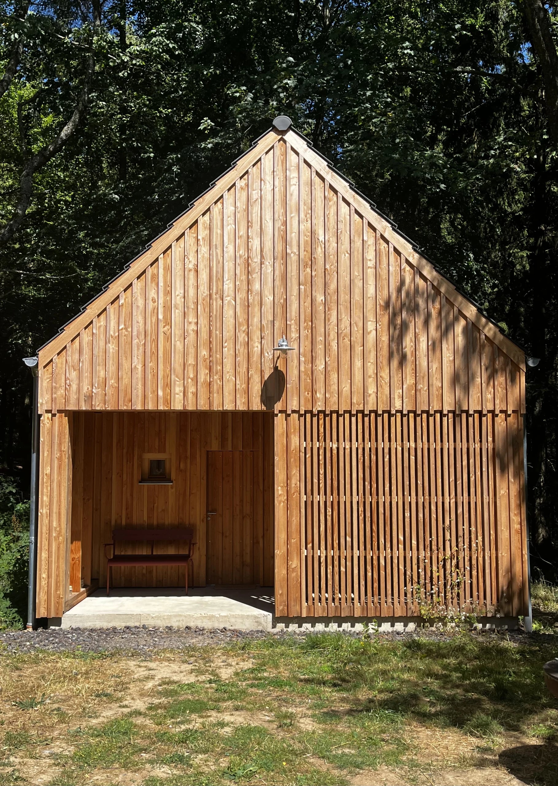 Die Saunahütte aus hellem Holz am Waldrand, davor eine überdachte Terrasse mit Bank und eine Außendusche.