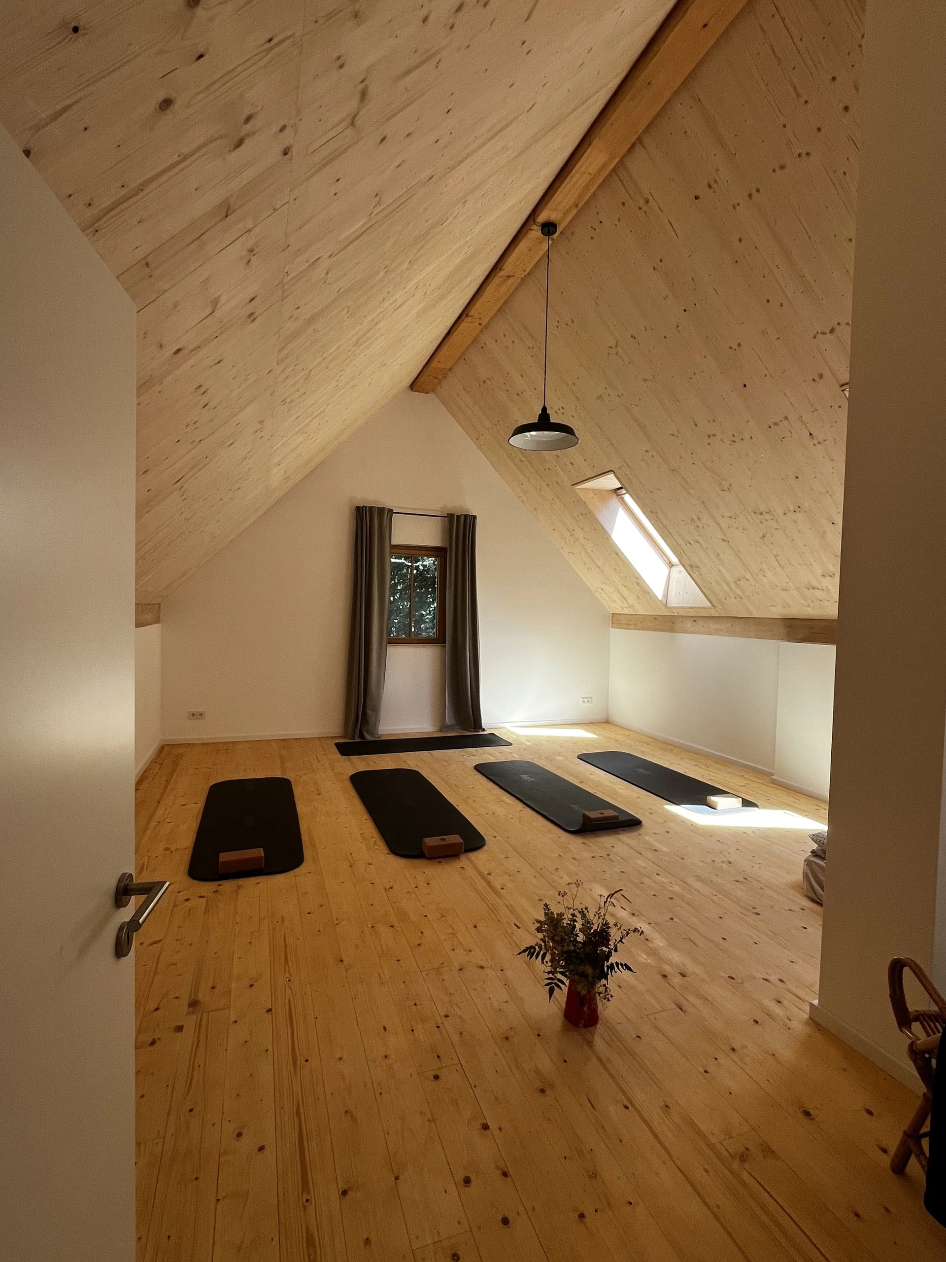 Der Yogaraum unter dem Dach mit Holzboden, Holzdecke und ausgelegten Matten, dazwischen eine Vase mit Wiesenblumen.