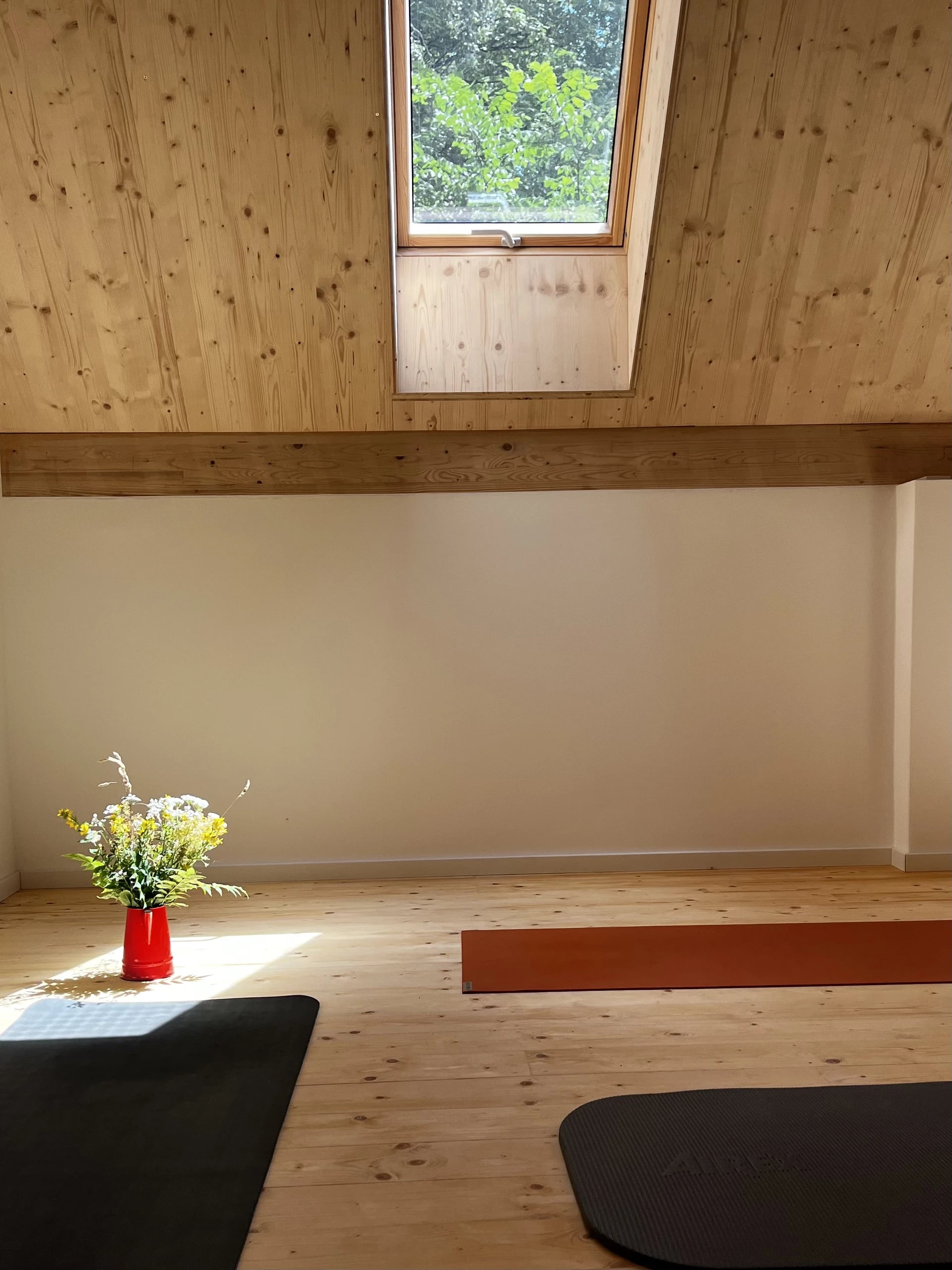 Morgenlicht fällt durch das Dachfenster des Yogaraums auf den Holzboden, daneben eine rote Vase mit Wiesenblumen.