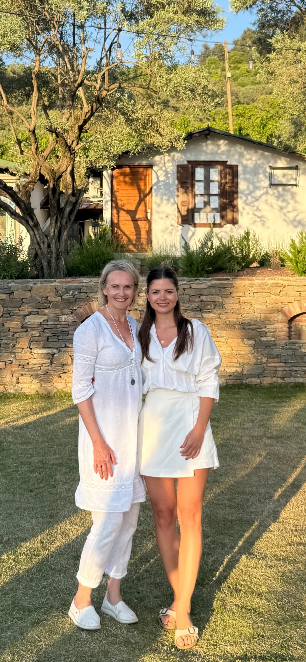 Astrid und Selin in Weiß stehen lächelnd im Garten vor einem Steinhaus im Abendlicht.