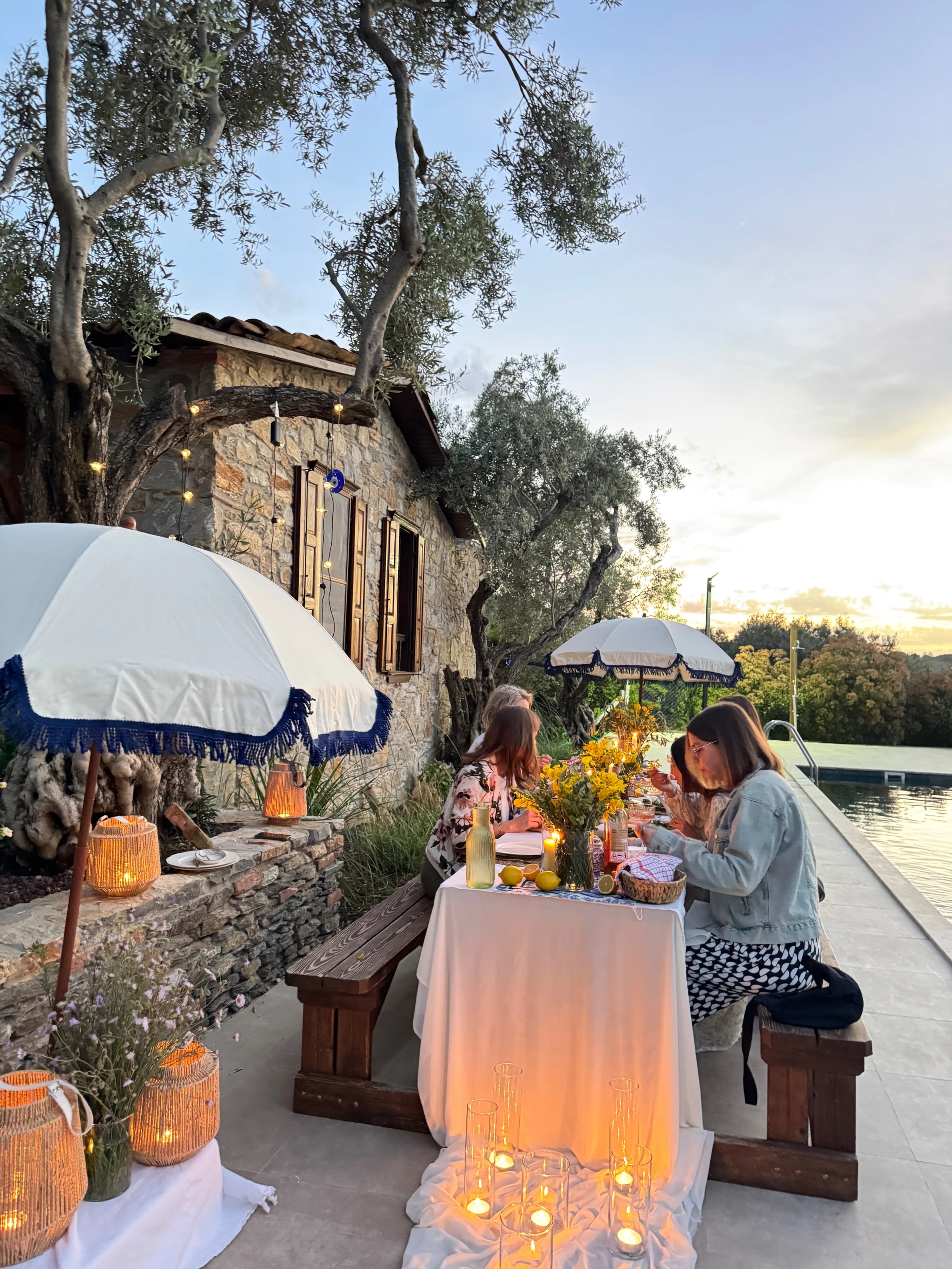 Die Gruppe sitzt abends an einer Tafel mit Kerzen und gelben Blumen am Pool vor einem Steinhaus.