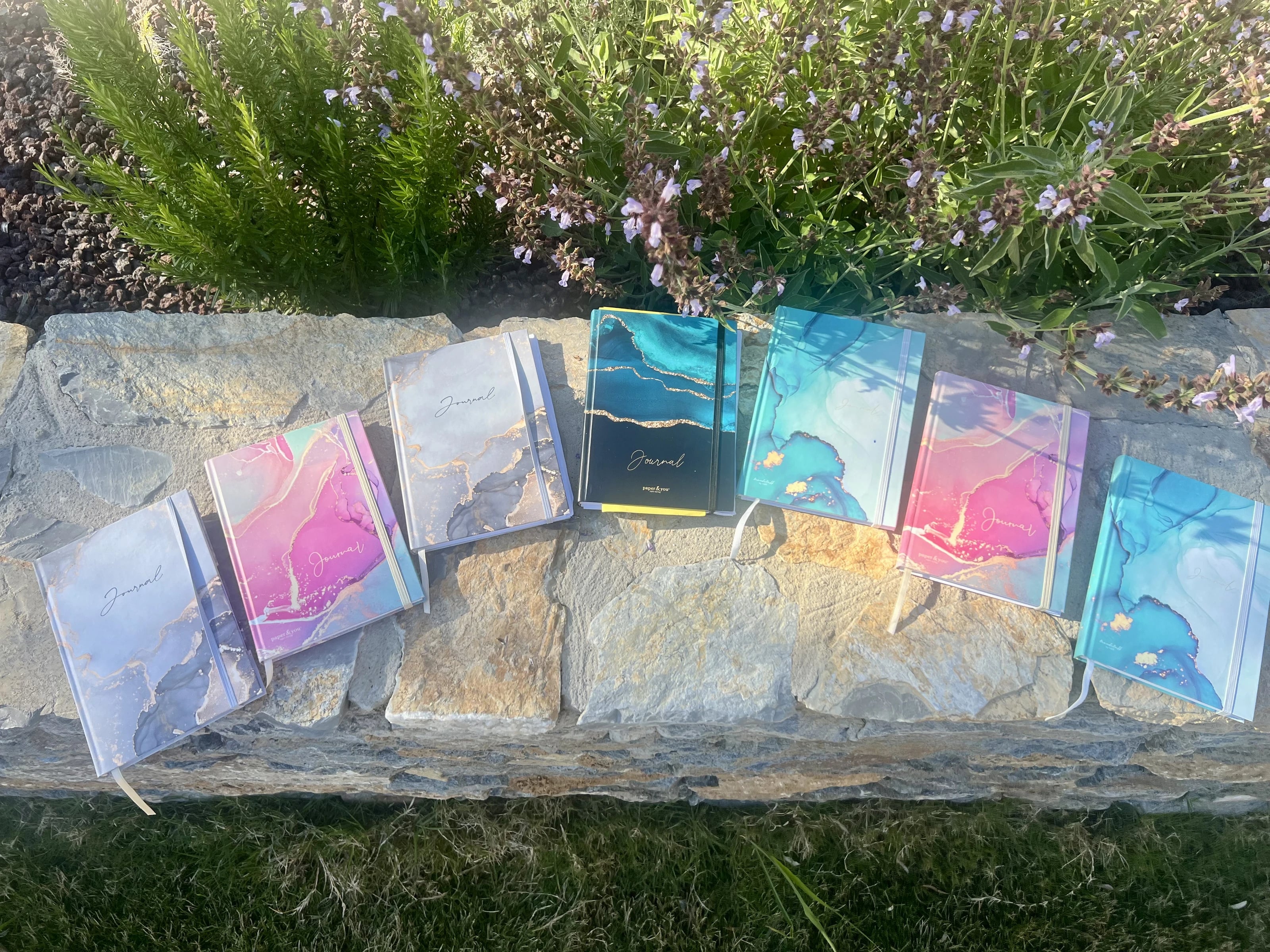 Bunte Journale liegen aufgereiht auf einer Steinmauer vor blühendem Salbei und Rosmarin.