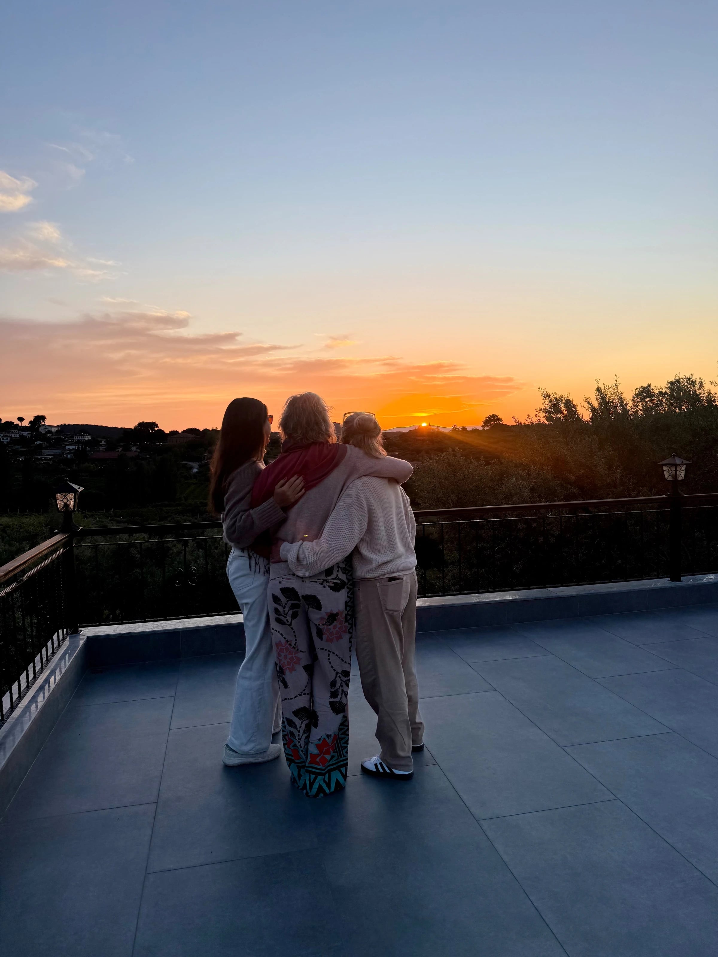 Drei Frauen stehen eng umarmt auf einer Terrasse und schauen in den Sonnenuntergang.