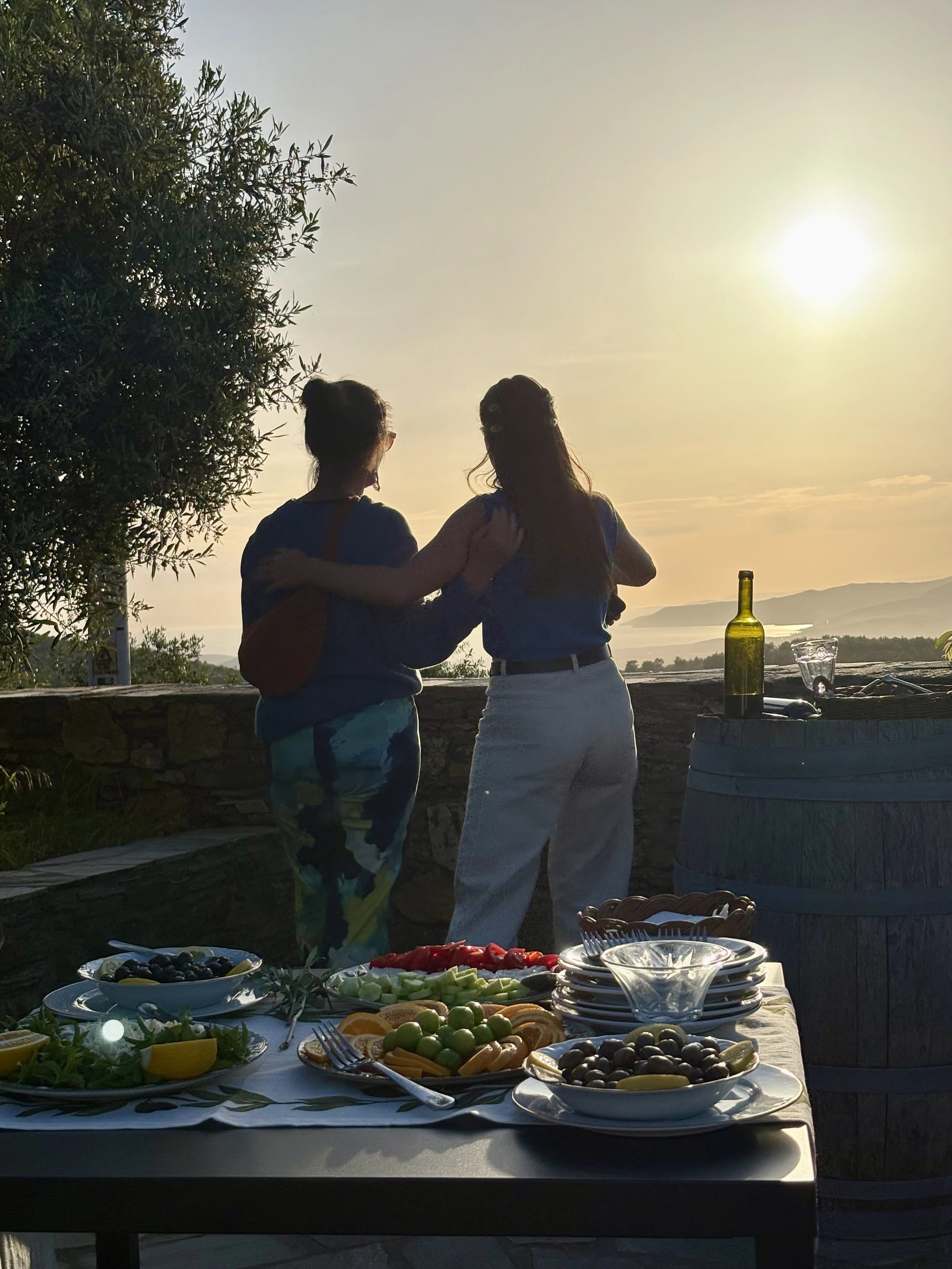 Zwei Frauen stehen Arm in Arm an einer Steinmauer und blicken in die tief stehende Sonne über dem Meer.