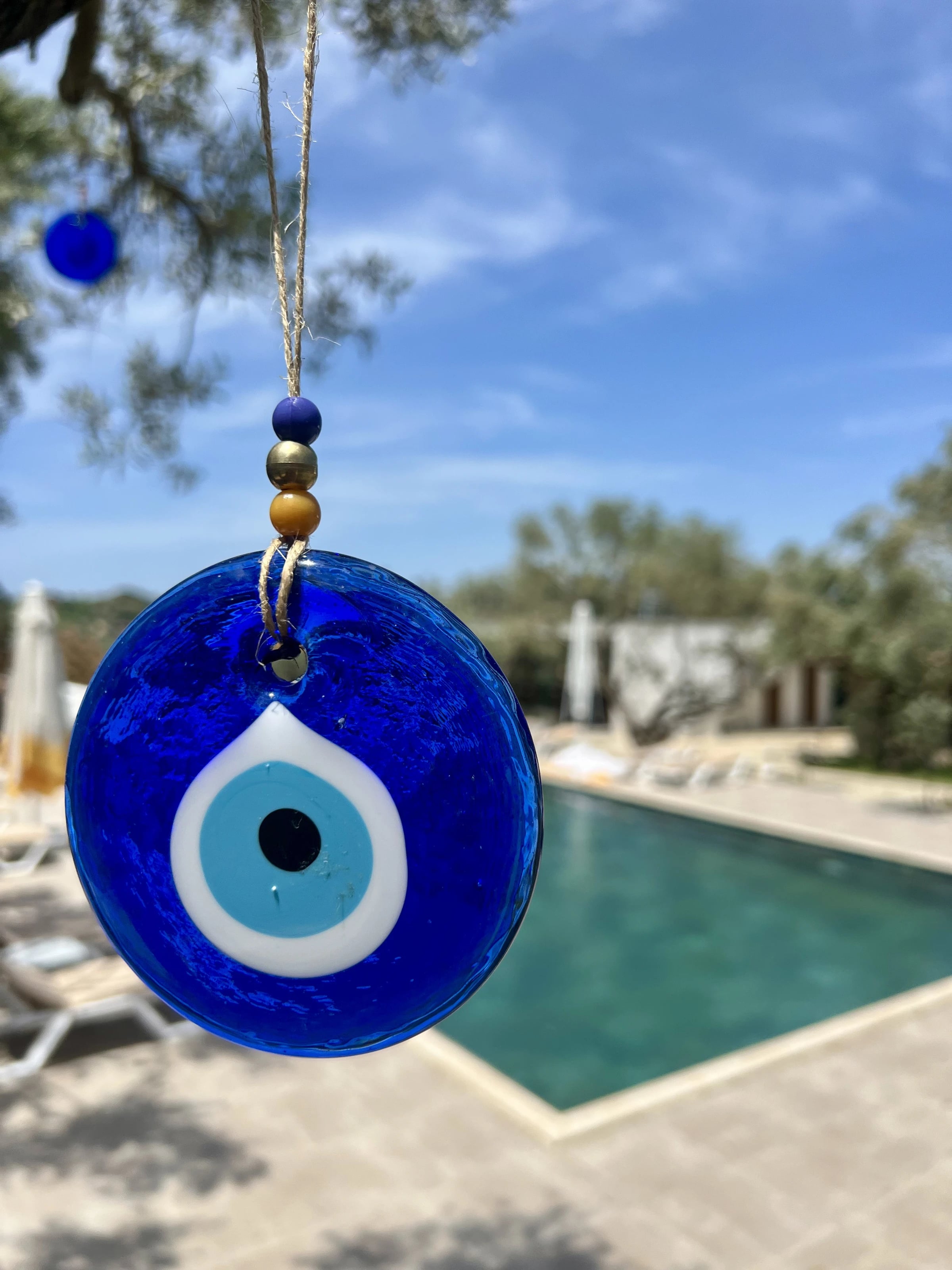 Ein blaues Nazar-Auge aus Glas hängt im Olivenbaum, dahinter unscharf der Pool.