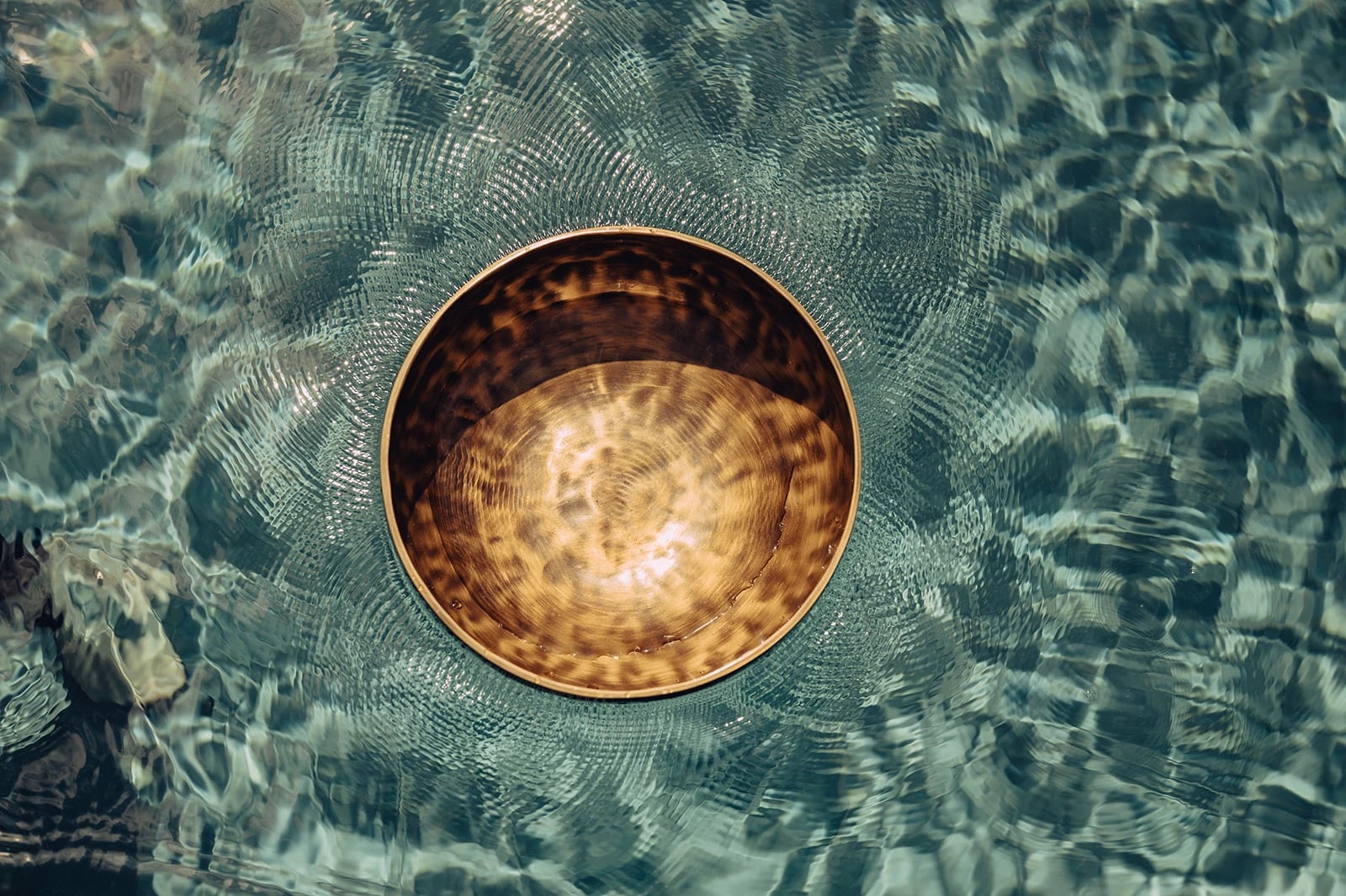 Eine goldene Klangschale schwimmt auf klarem, türkisfarbenem Wasser, um sie herum feine Wellenringe.