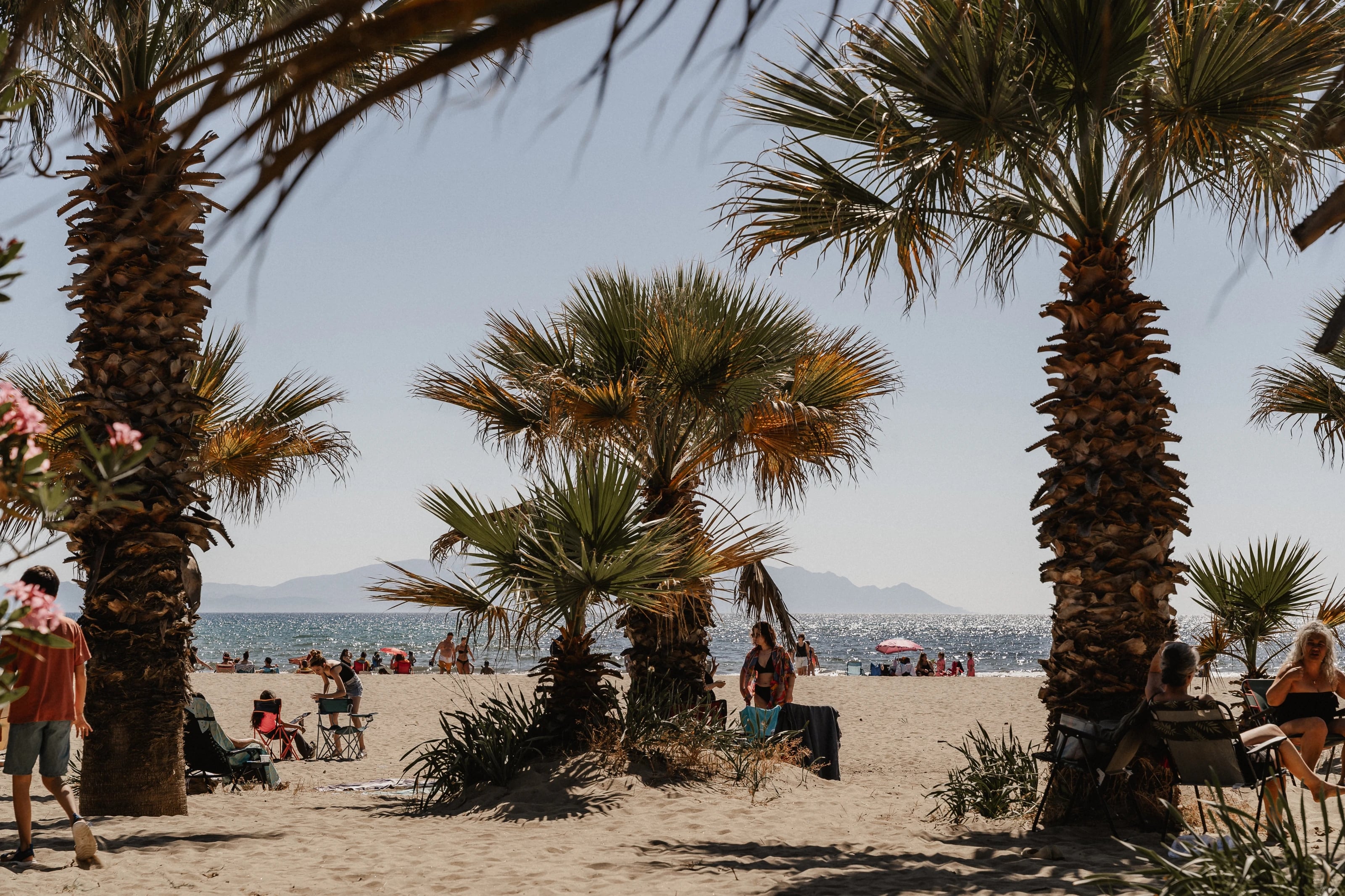 Sonniger Sandstrand mit Palmen, dahinter das glitzernde Meer und Berge am Horizont.
