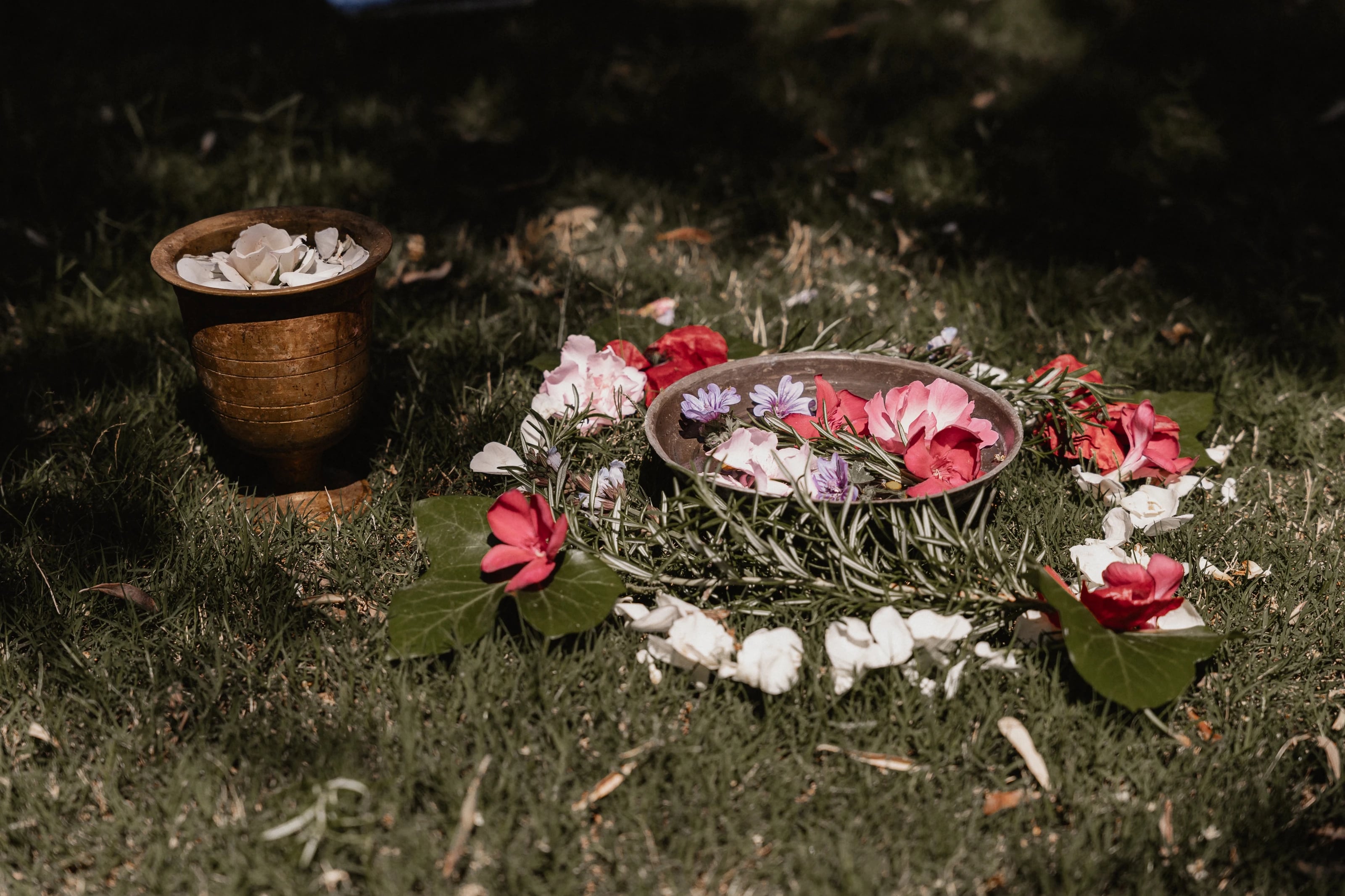 Ritualplatz im Gras mit einer Schale voller Blüten, Rosmarinzweigen und einem Messingtopf mit Blütenblättern.