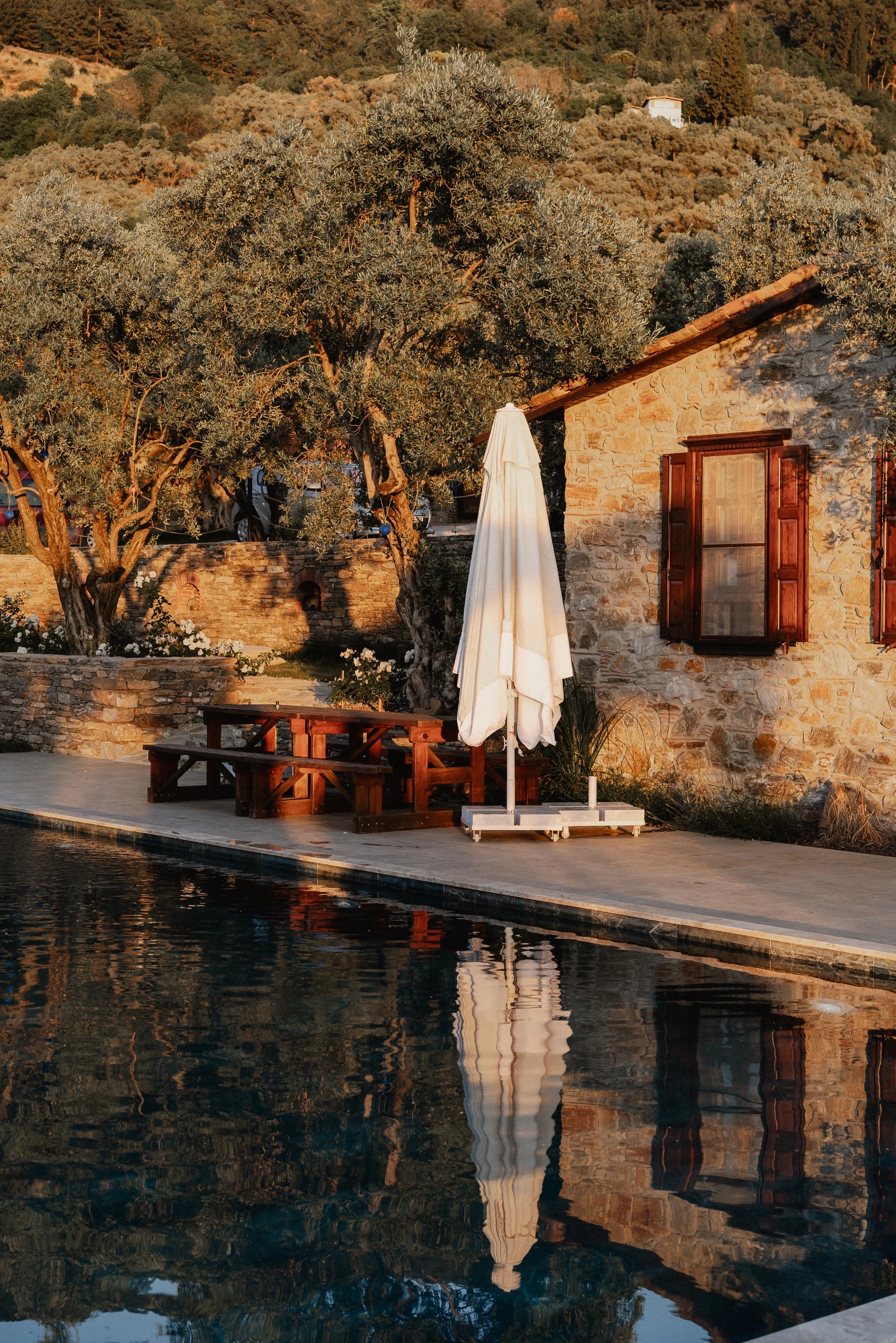 Abendsonne auf dem Steinhaus am Pool, davor ein geschlossener Sonnenschirm und Holzbänke, im Wasser die Spiegelung.