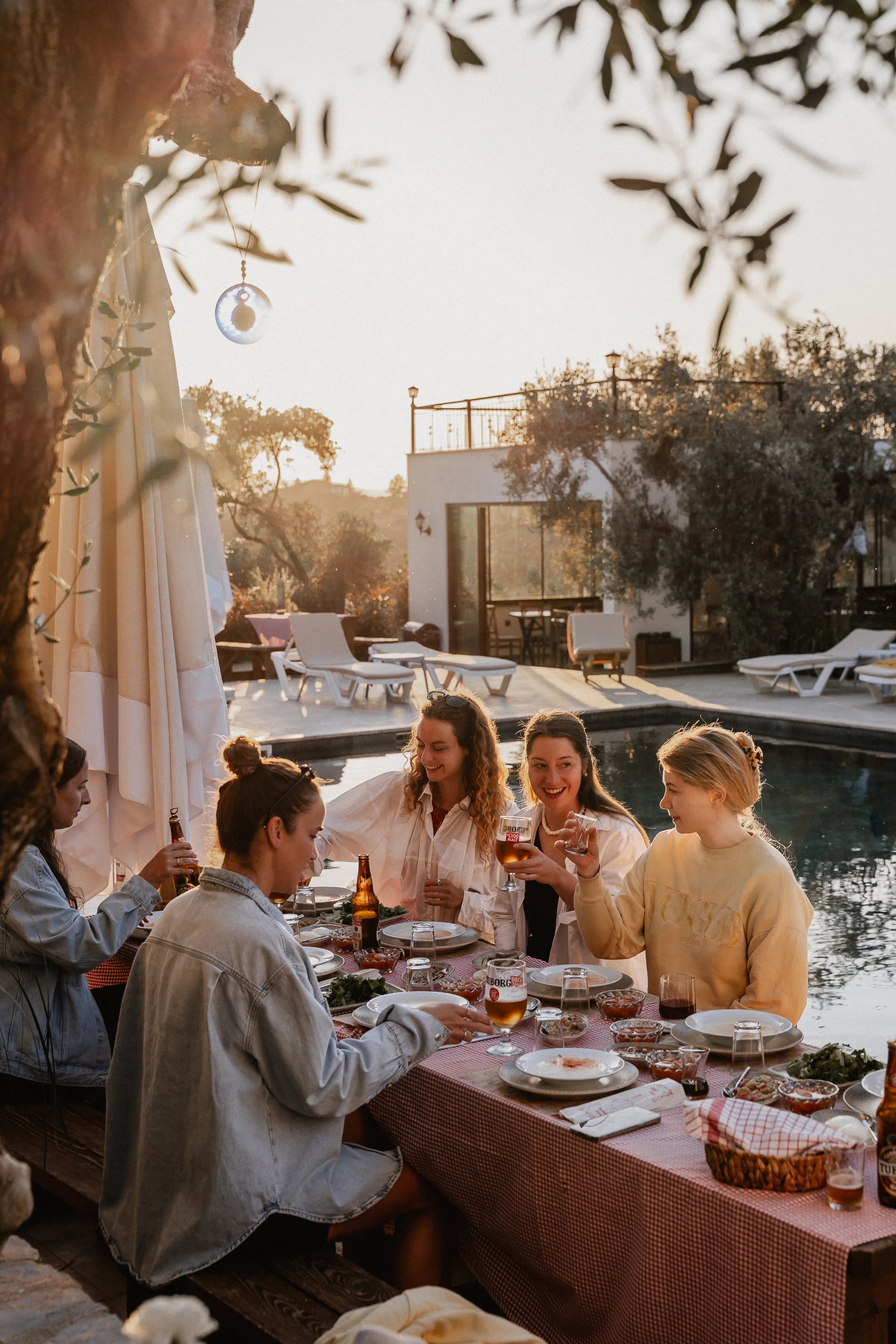 Gruppe Frauen sitzt lachend beim Abendessen am Pool bei Sonnenuntergang