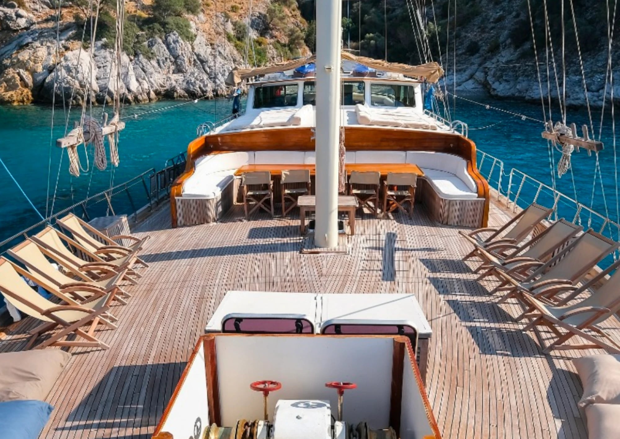 Das Holzdeck der Larin Su mit Liegestühlen und Sitzecke, vor Anker in einer Bucht mit türkisfarbenem Wasser und Felsen