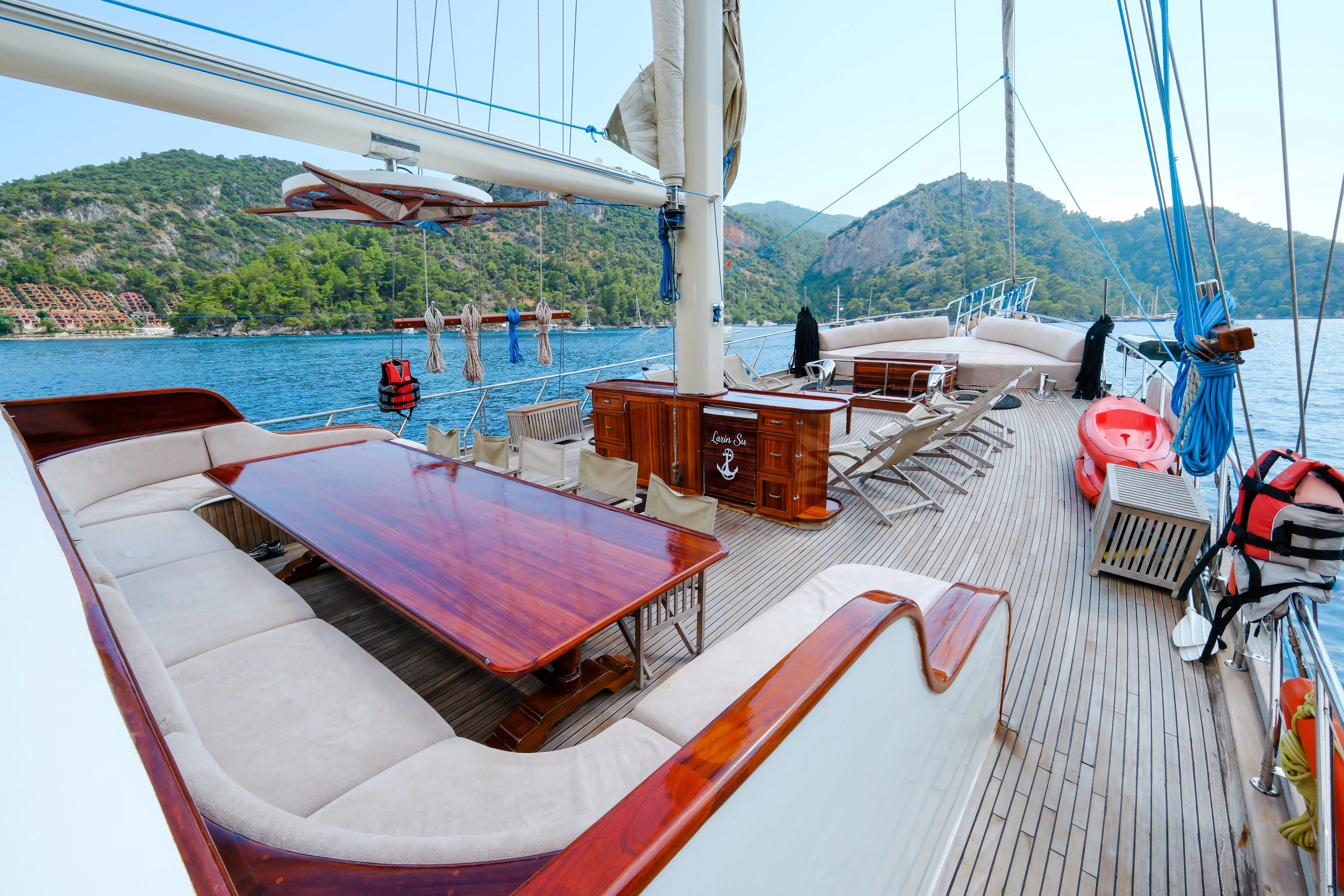 Das geräumige Deck der Larin Su mit Sitzecke, Holztisch und Liegestühlen vor bewaldeten Hügeln.