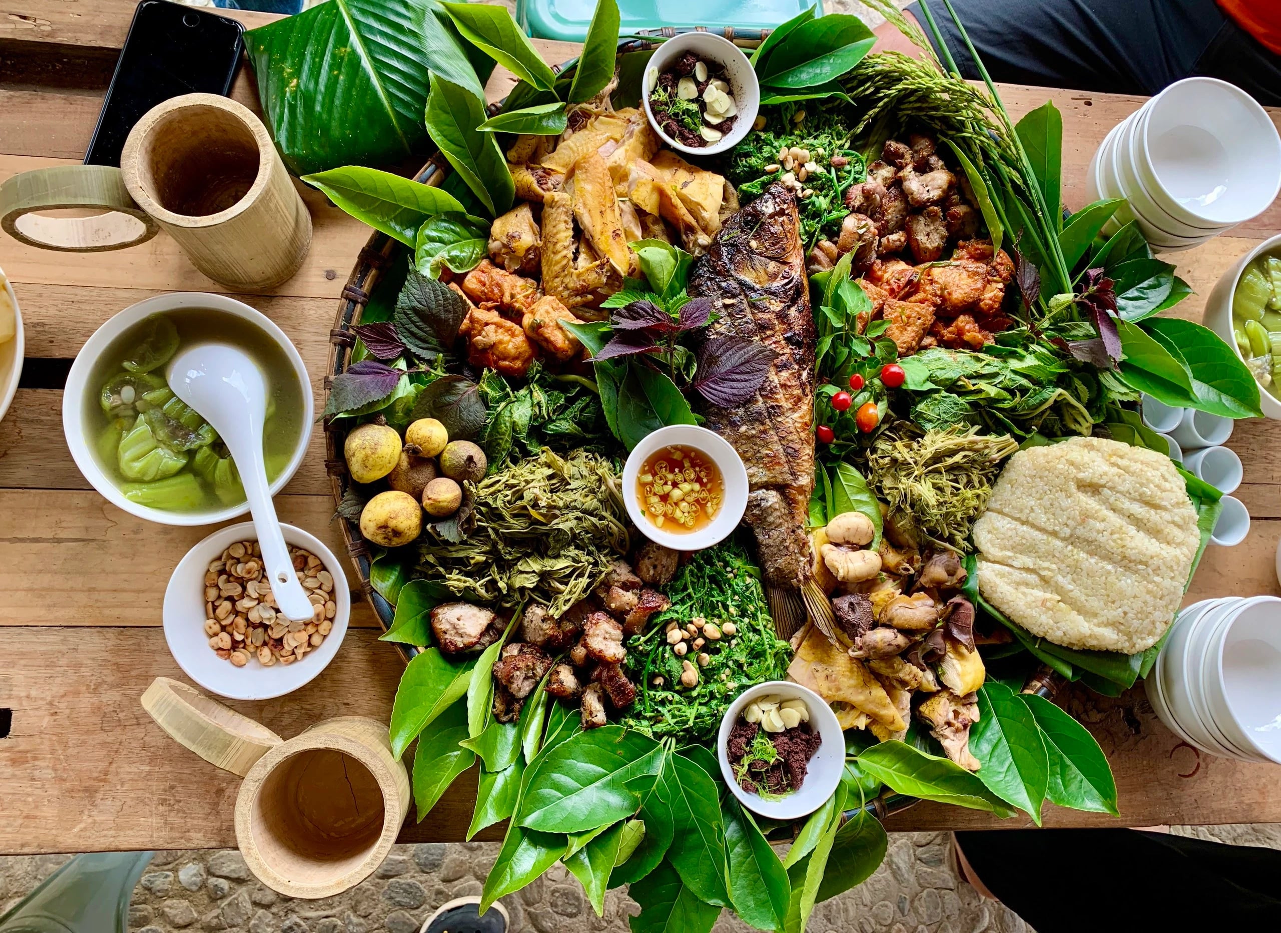 Eine große Bambusplatte auf Bananenblättern mit gegrilltem Fisch, Hühnchen, Wildkräutern, Klebreis und kleinen Saucenschalen, dazu Becher aus Bambus.