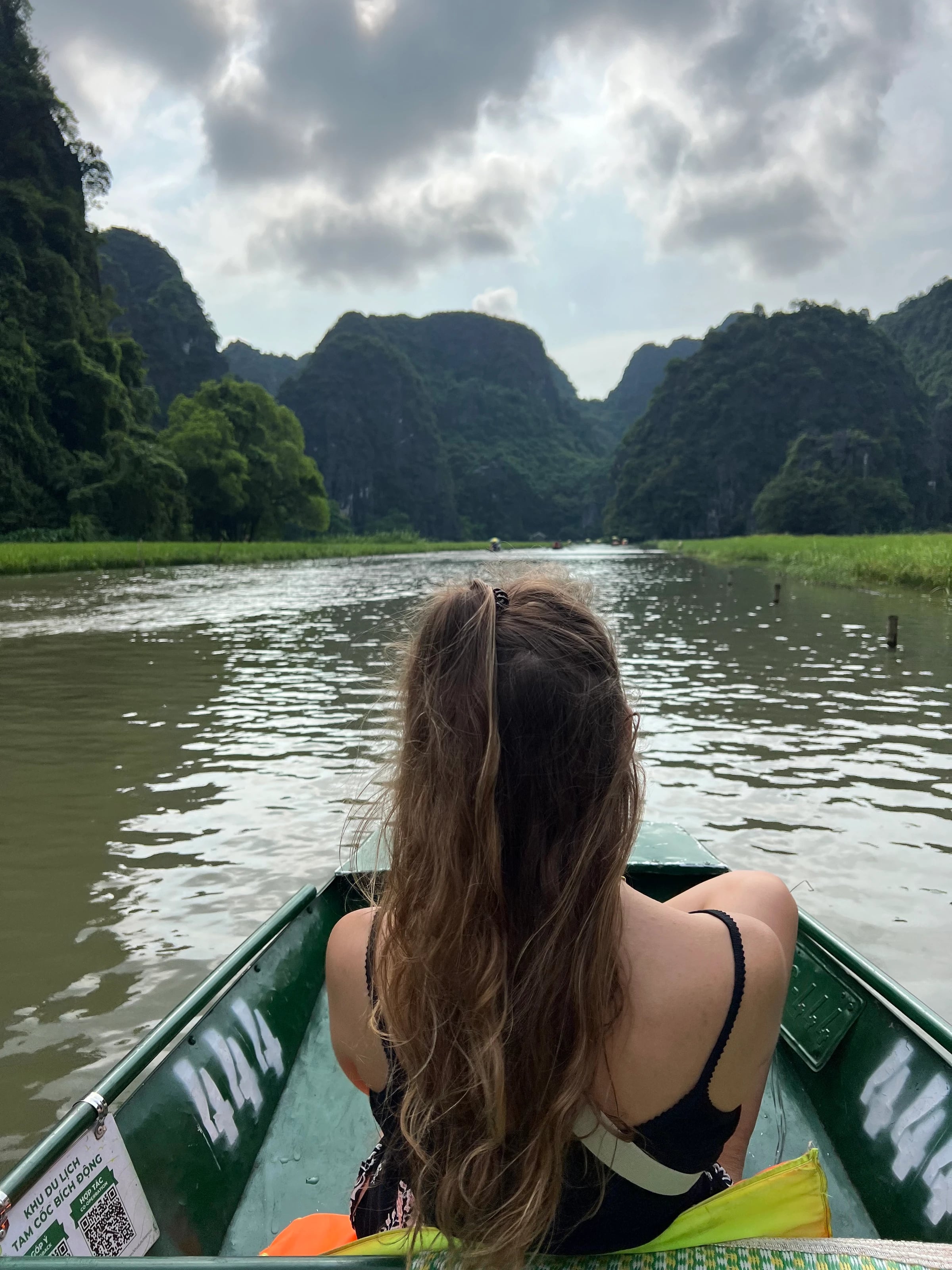 Eine Frau sitzt im Ruderboot und blickt über den Fluss auf die Karstberge von Tam Coc.