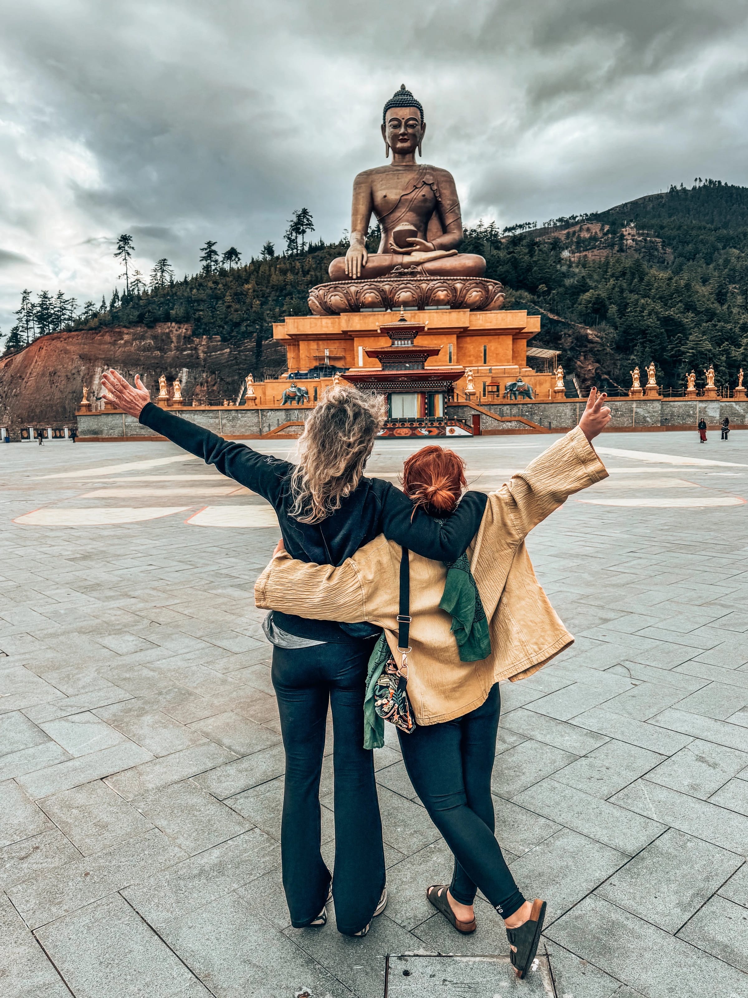 Zwei Reisende breiten die Arme aus vor der großen goldenen Buddha-Dordenma-Statue.