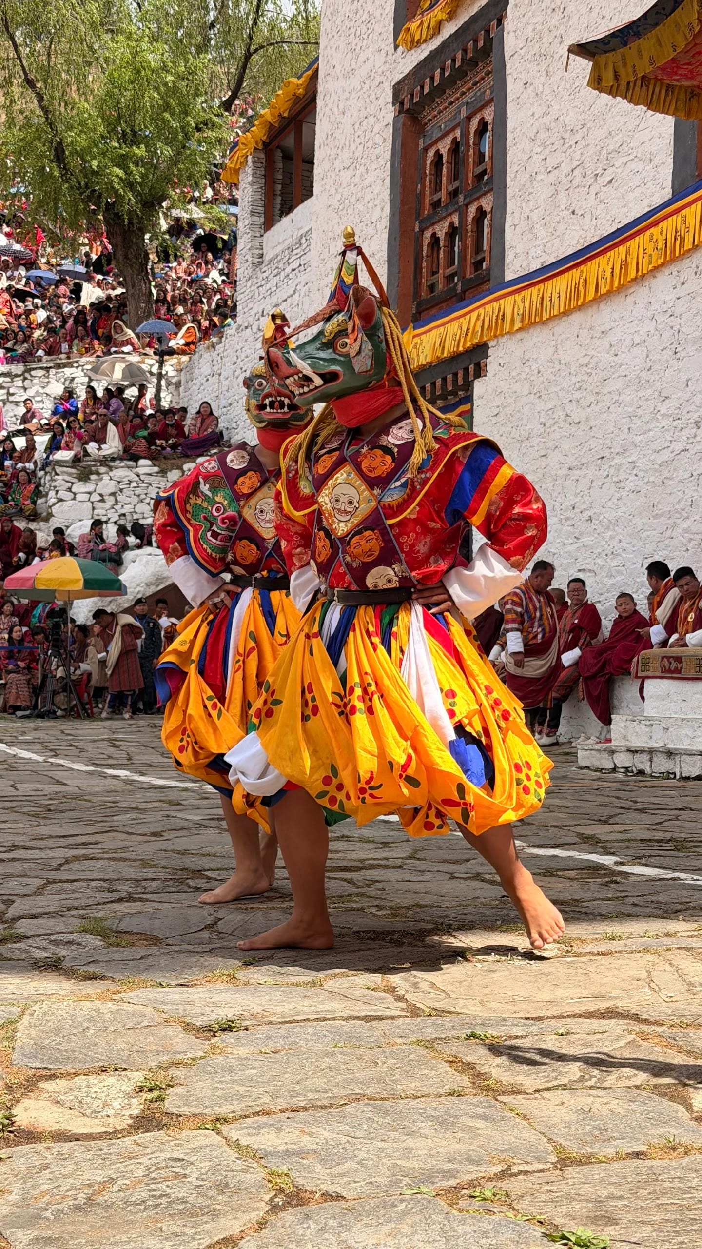 Maskentänzer in bunten Seidengewändern mit gelben Röcken und Tiermasken tanzen barfuß auf dem Steinhof eines Klosters in Bhutan, am Rand sitzen dicht gedrängt die Zuschauer.