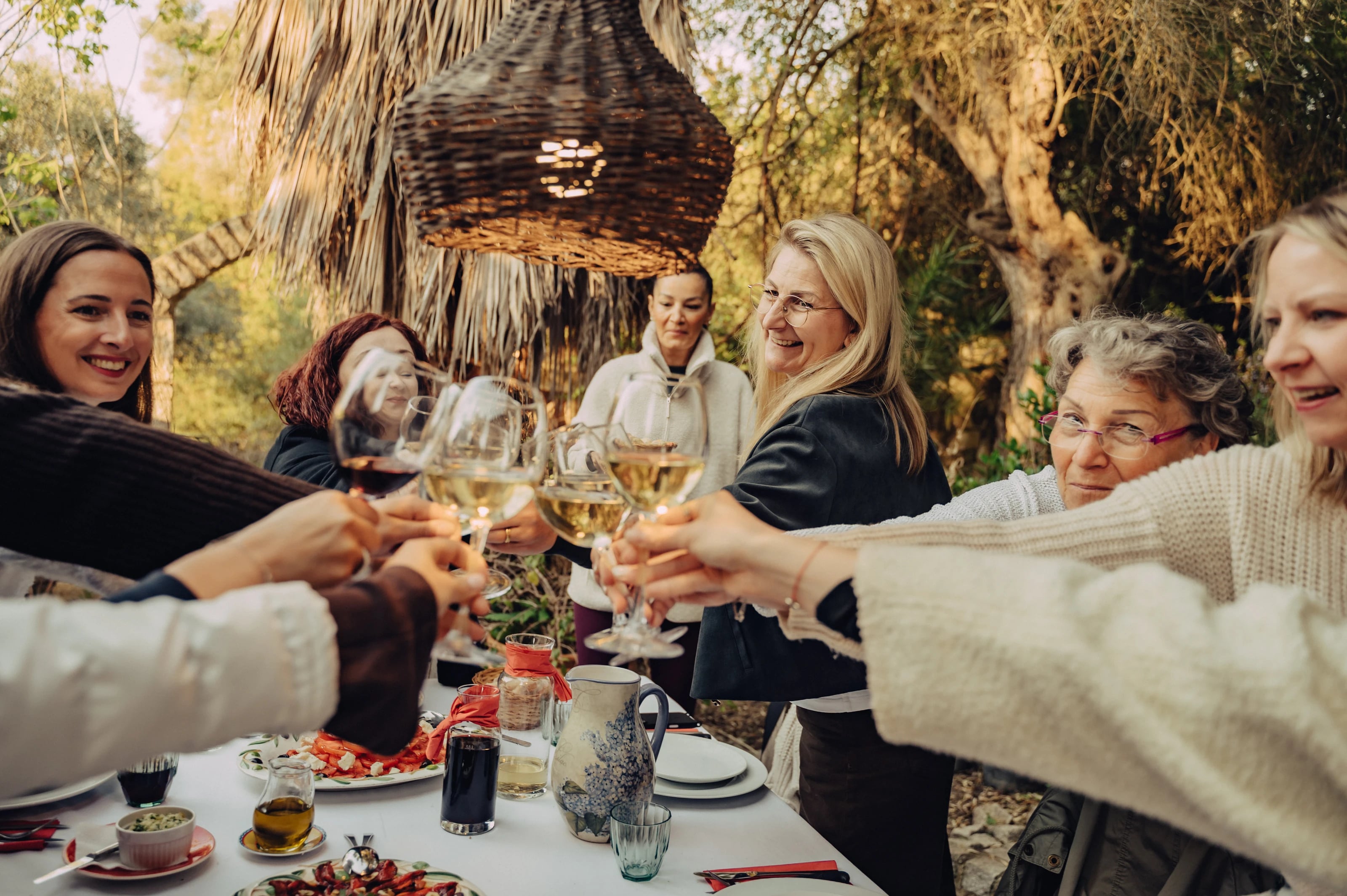 Eine Gruppe Frauen stößt lachend mit Weingläsern an einer gedeckten Tafel im Freien an.