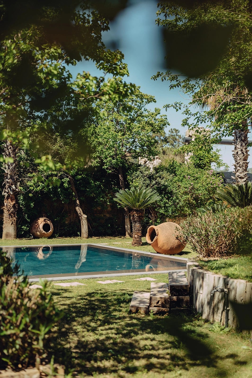 Blick durch Blätter auf einen Pool im grünen Garten mit Palmen und großen Tonkrügen.