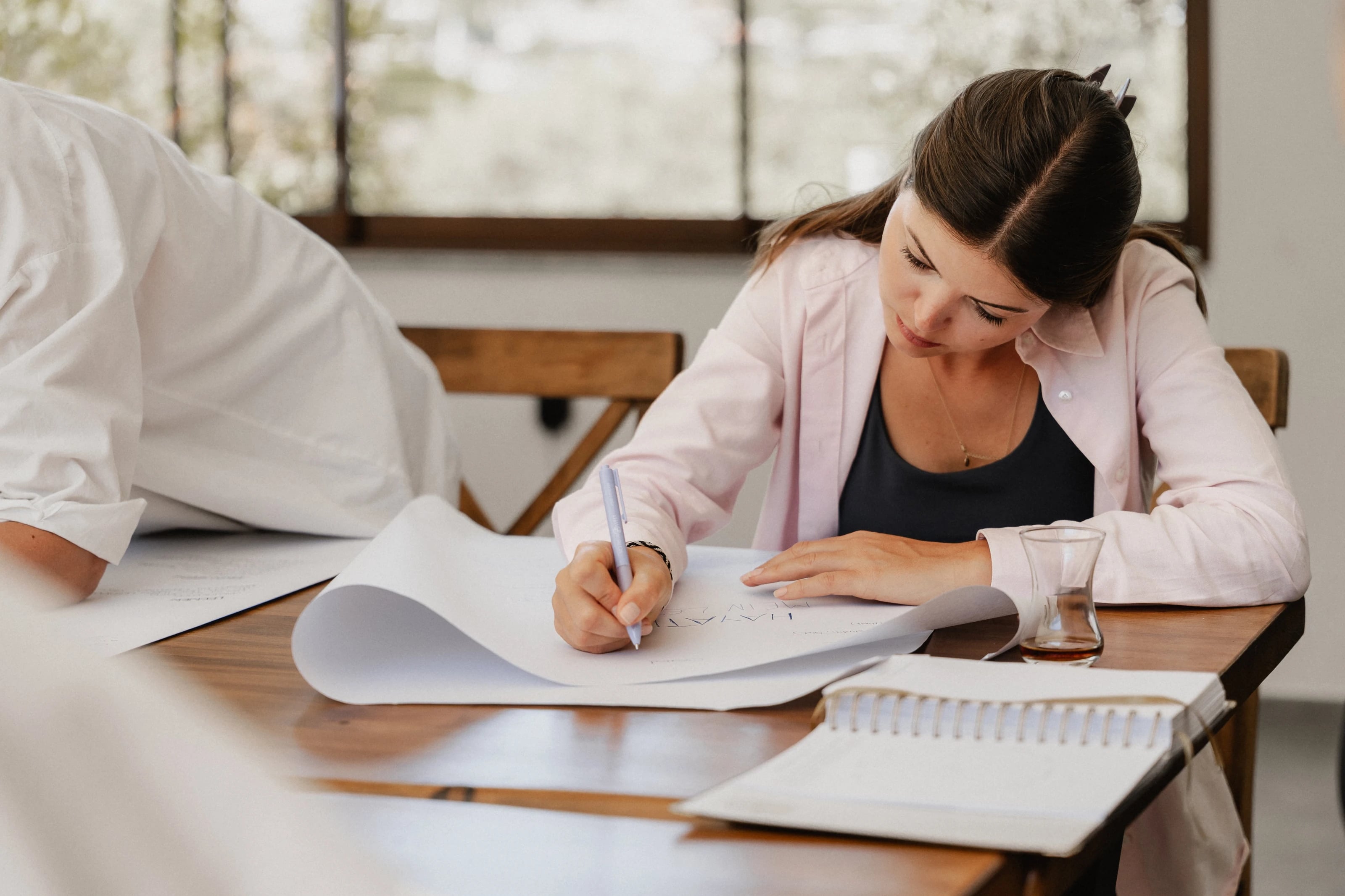 Eine Frau macht sich Notizen auf einem Block am Holztisch, im Hintergrund eine weitere Person.
