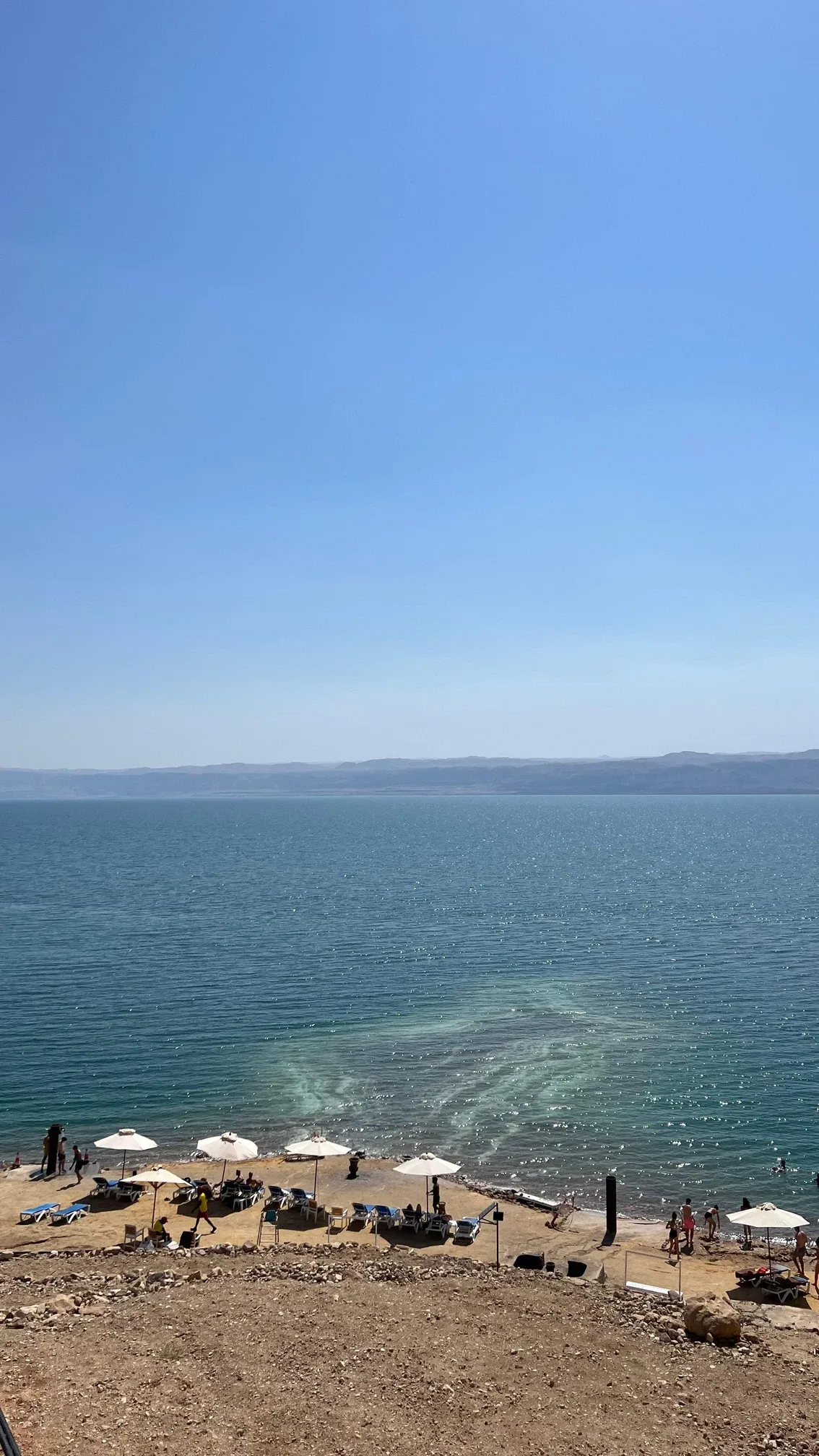 Blick von oben auf das türkisfarbene Tote Meer mit Liegestühlen und Sonnenschirmen am Ufer und Hügeln in der Ferne.