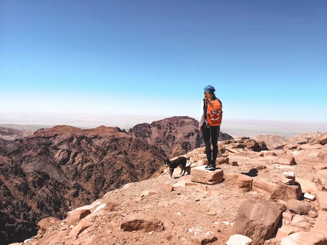 Eine Frau mit orangefarbenem Rucksack steht auf einem felsigen Berggipfel neben einem Hund mit Blick auf eine rote Bergkette.