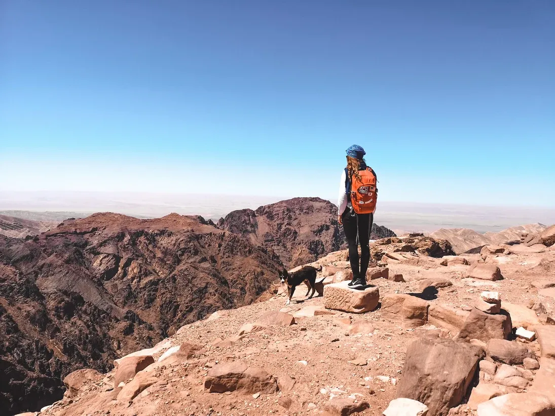 Eine Frau mit orangefarbenem Rucksack steht auf einem felsigen Berggipfel neben einem Hund mit Blick auf eine rote Bergkette.