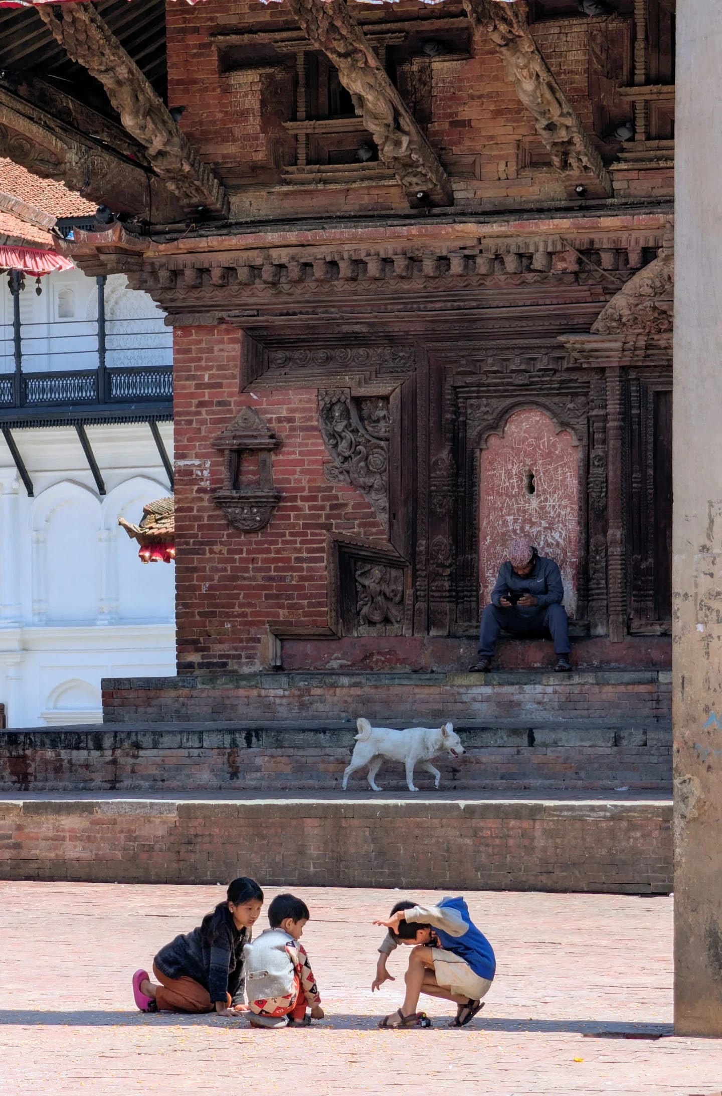 Drei Kinder hocken spielend auf einem Platz vor einem Backsteintempel mit geschnitzten Holzfenstern in Kathmandu, auf den Stufen läuft ein weißer Hund vorbei.